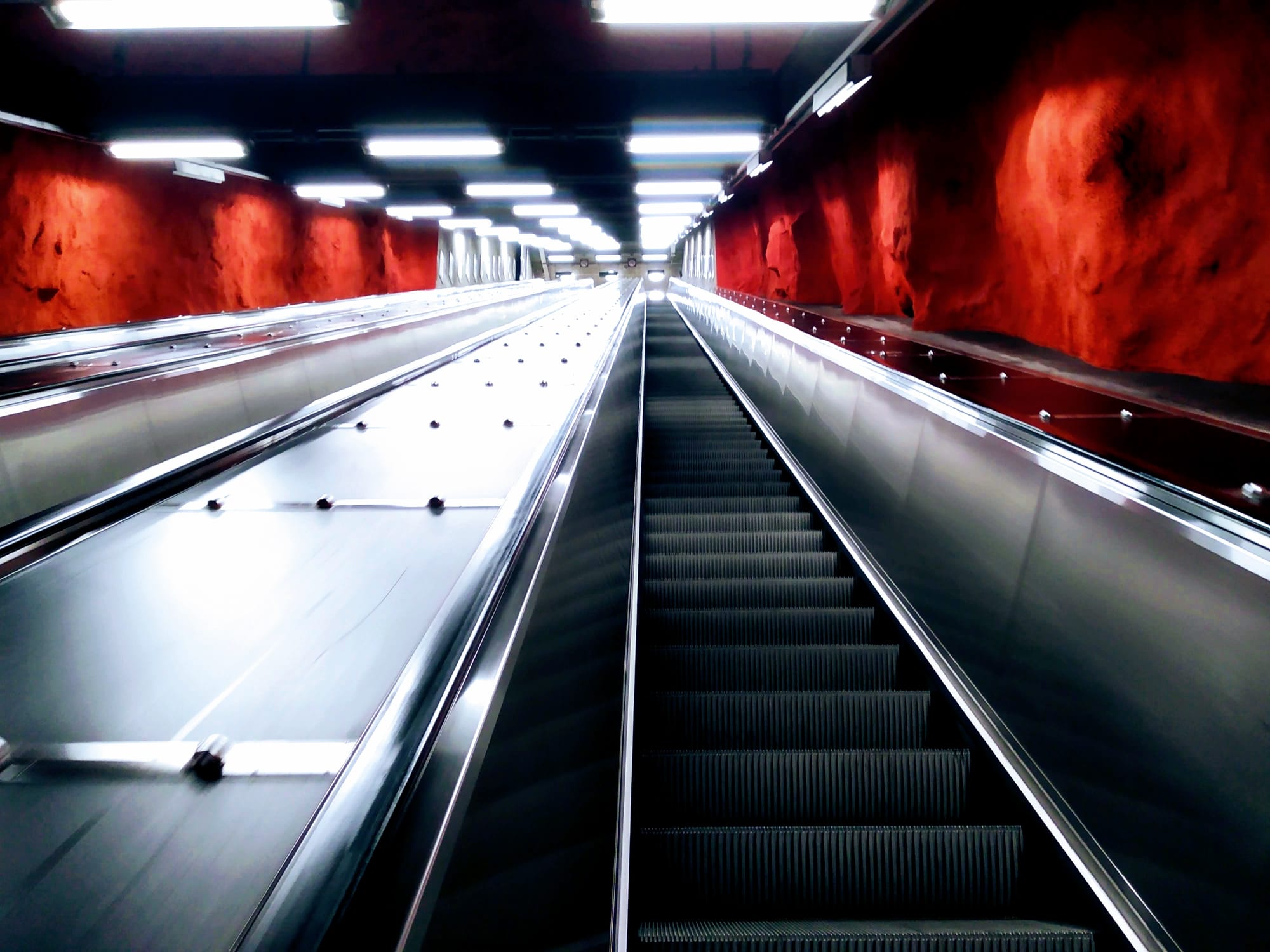 A silver escalator underneath bright white lights rises through red walls.