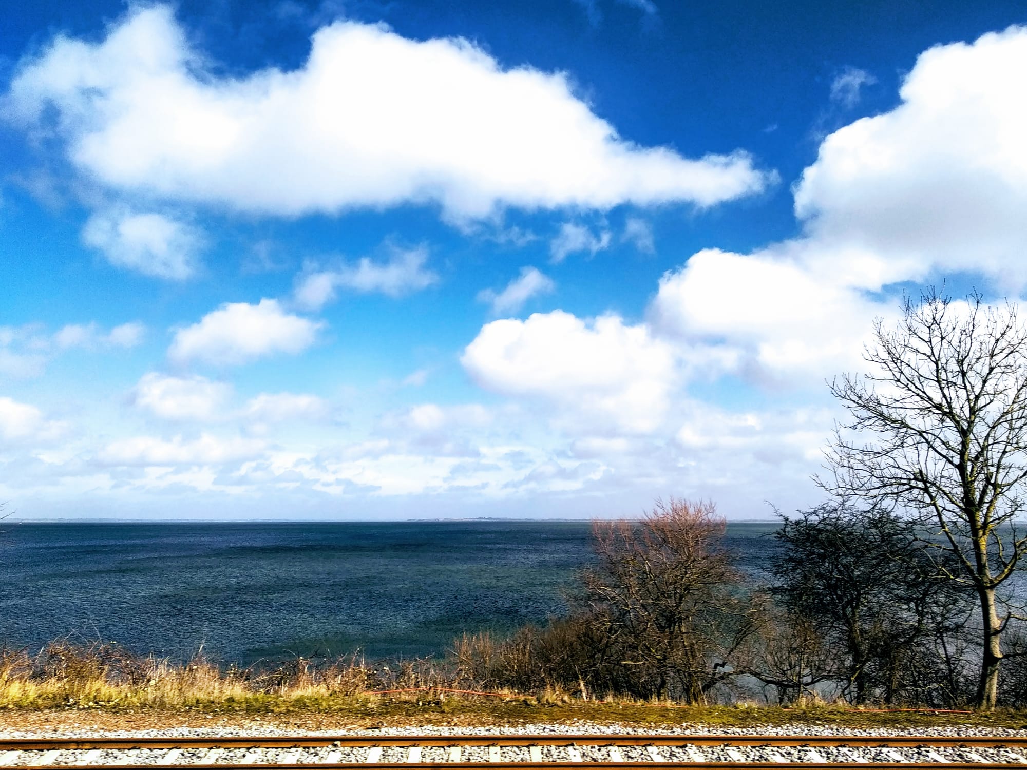 Little fluffy clouds hang in the sky above the water in Venø Bay. A train track runs along the foreground of the image.