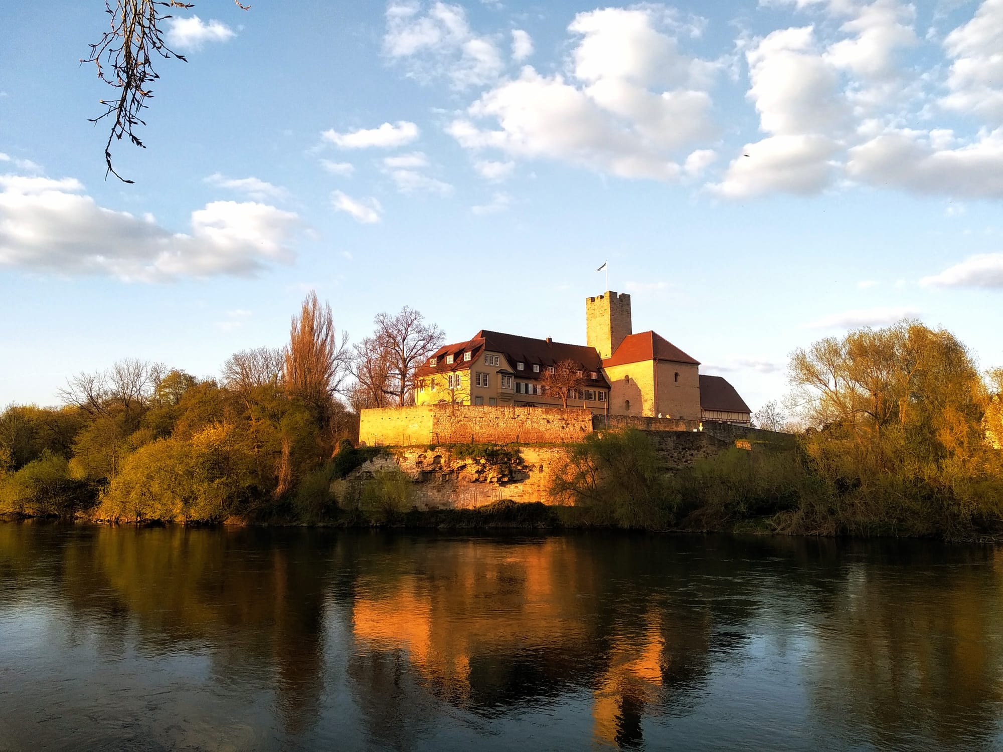 The fortified Lauffen city hall against a blue sky, surrounded by trees and reflected in the river Nekkar. 