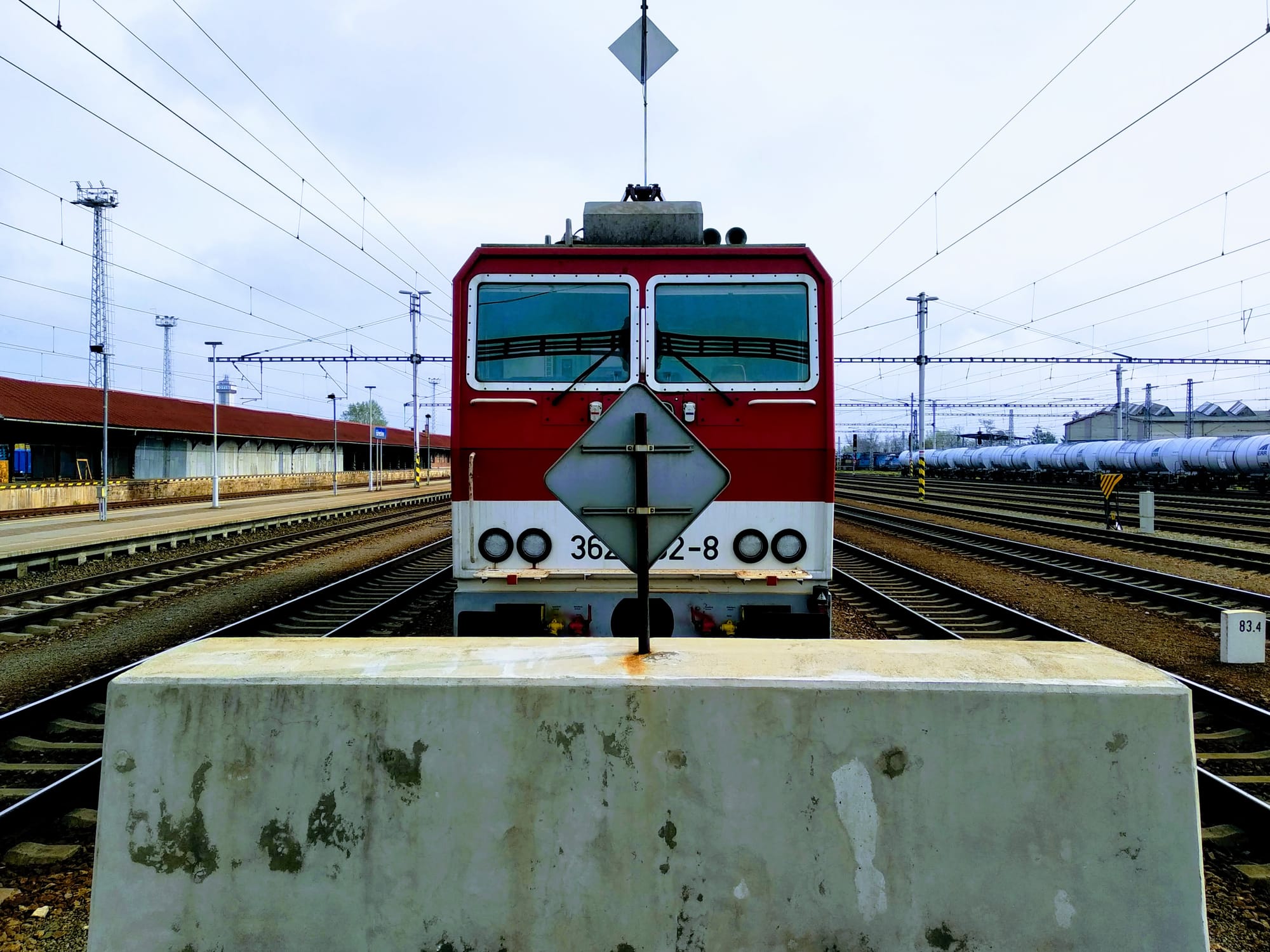 A red and white Czech locomotive parked at the end of a platform with train tracks either side converging behind it.