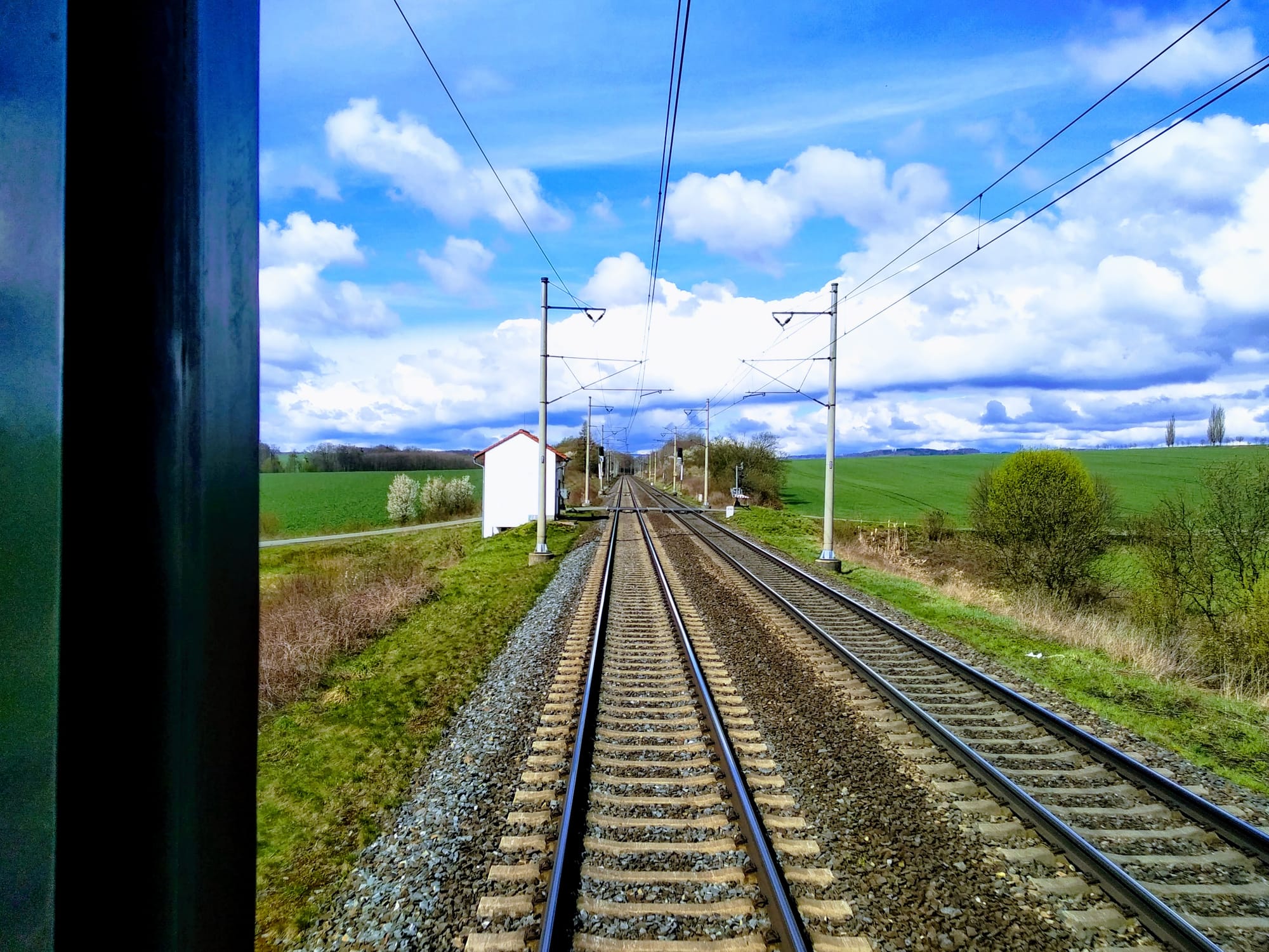 Seen from the back of a train, between green fields two sets of train tracks converge on the horizon under cumulus clouds. 