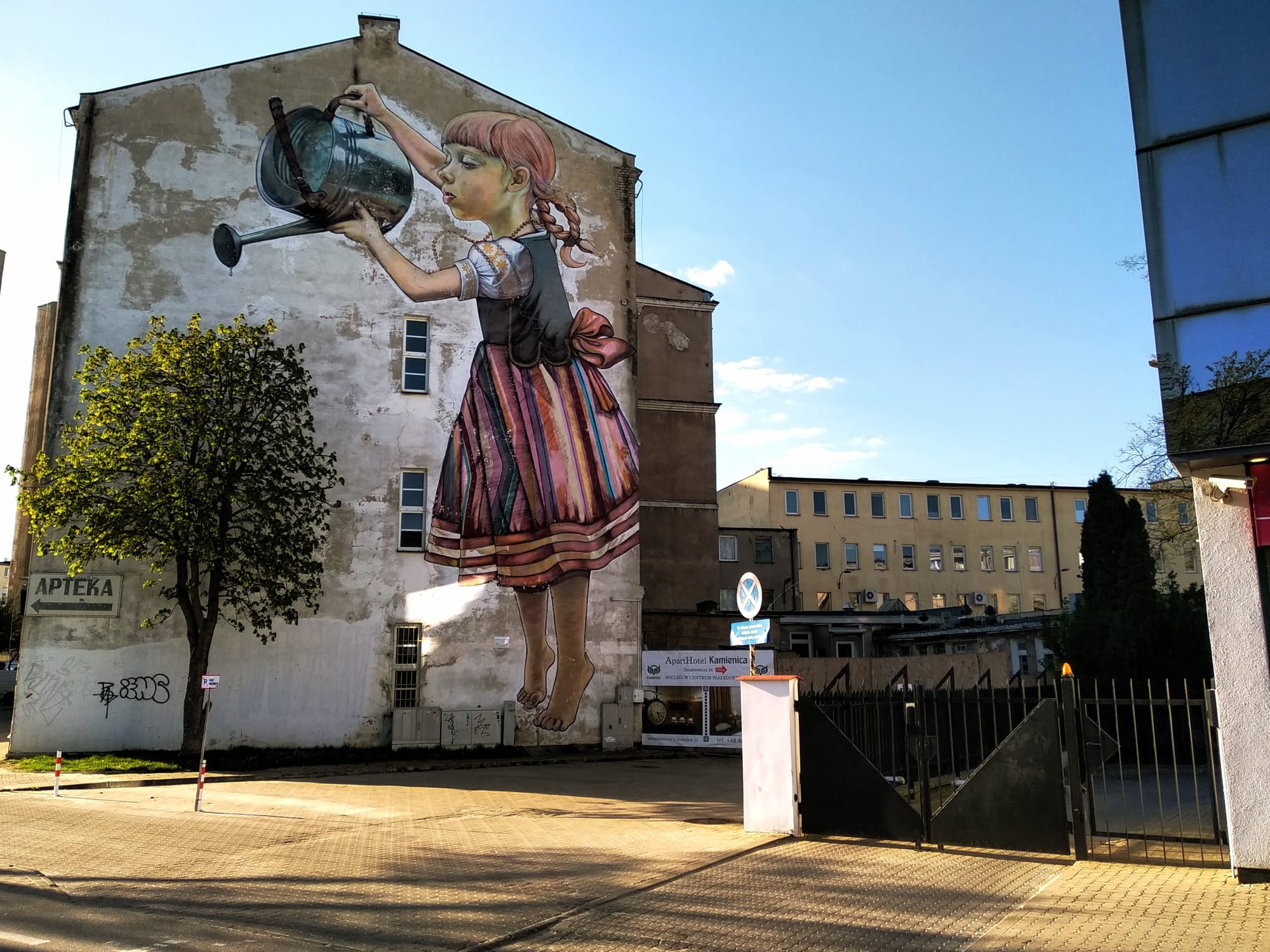 A young girl standing on tip-toes holds a watering can painted onto the gable end of a building which she uses to water a tree planted in front of the wall.