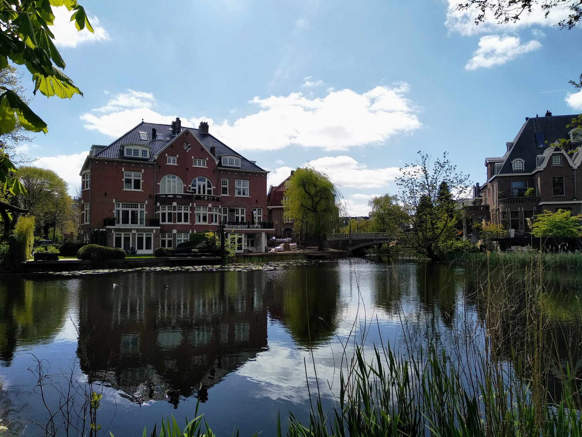Blue sky, clouds, trees, and large red brick houses reflect in water in a park in Amsterdam.
