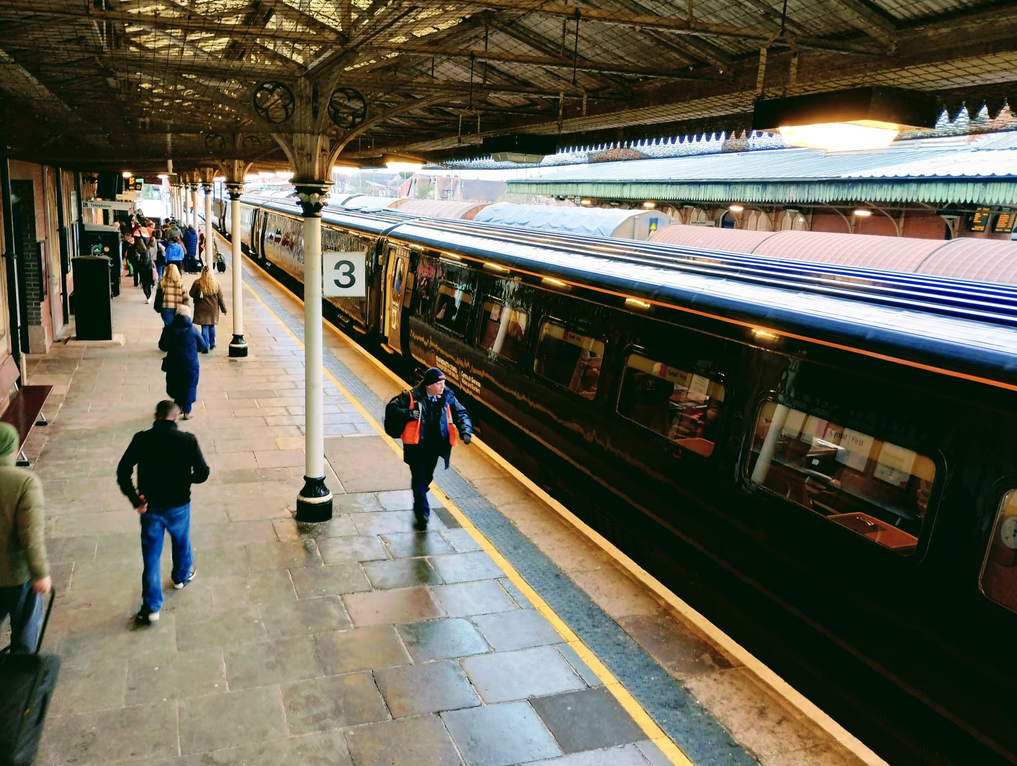 A Transport for Wales train waiting on platform 3 in Hereford train station. People walk along the platform away from the camera. A rail worker in a hi-visibility jacket is walking towards us.