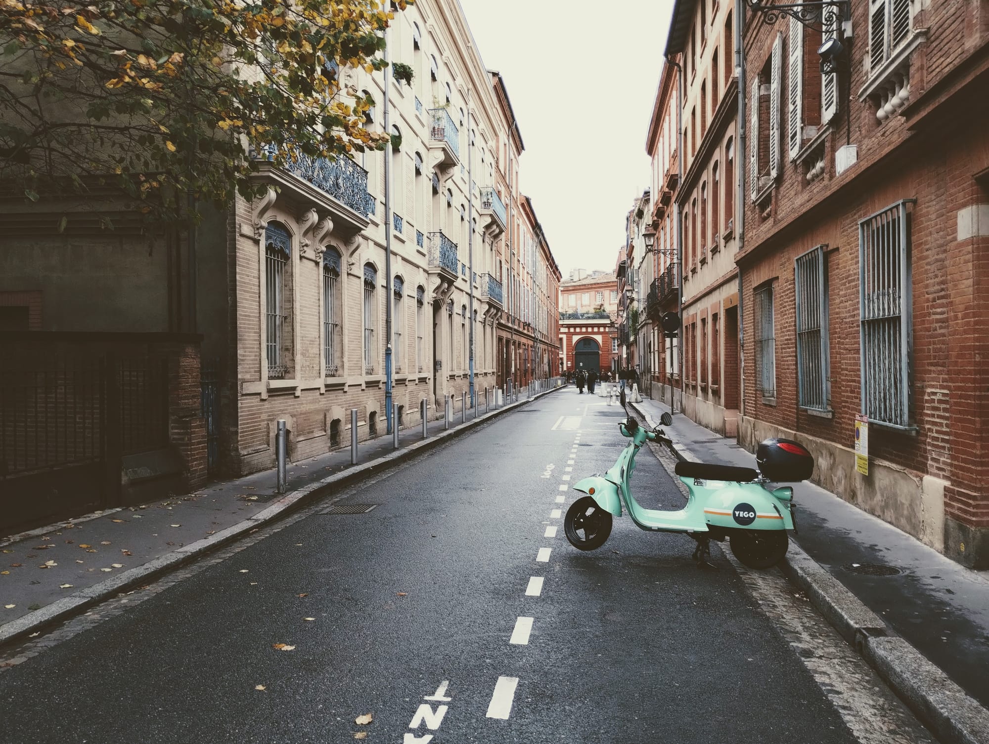 A baby-blue moped stands at one end of a slightly damp street flanked by brick buildings.