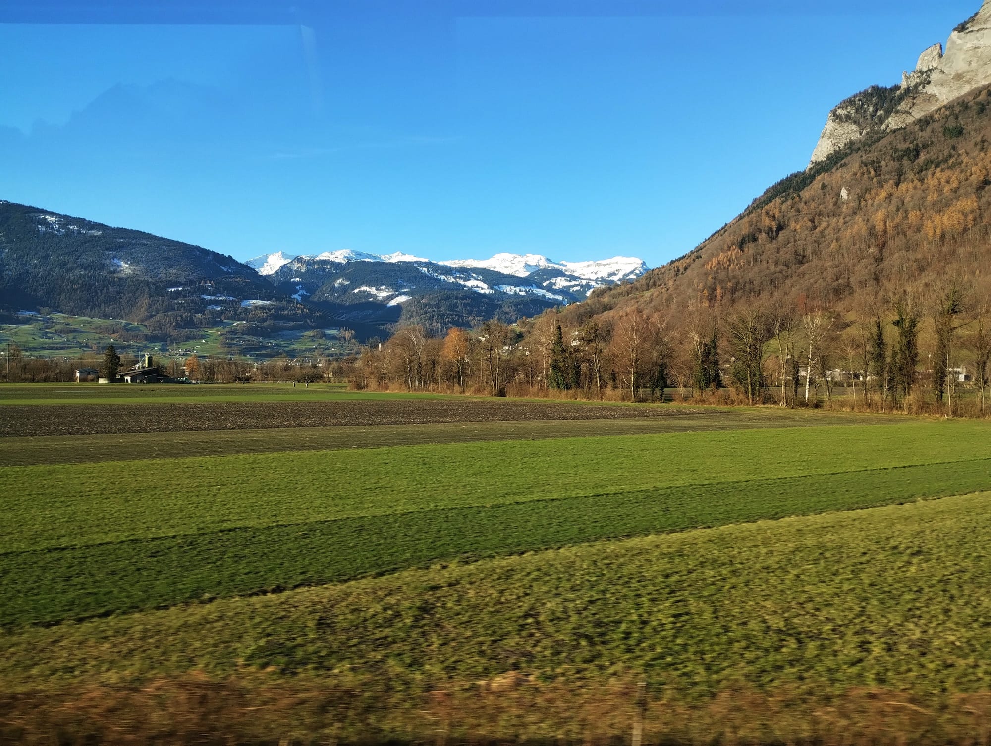 Snow-capped mountains dominate the horizon behind green fields and woodland.