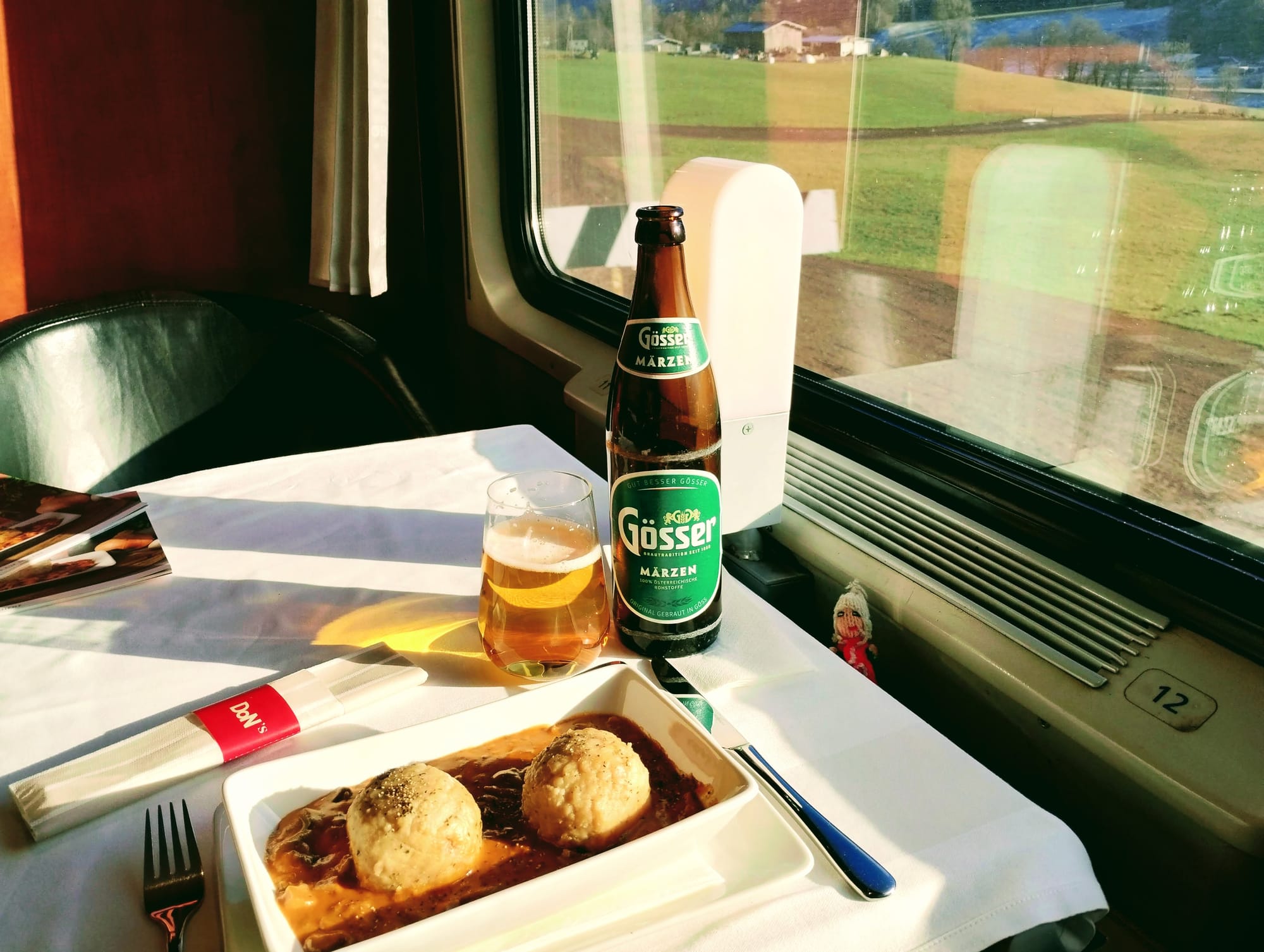 In a dining car next to a window, dumplings and mushroom sauce served in a china dish with a glass and a bottle of Gösser. A Peruvian finger puppet enjoys the view. Fields pass by the window.