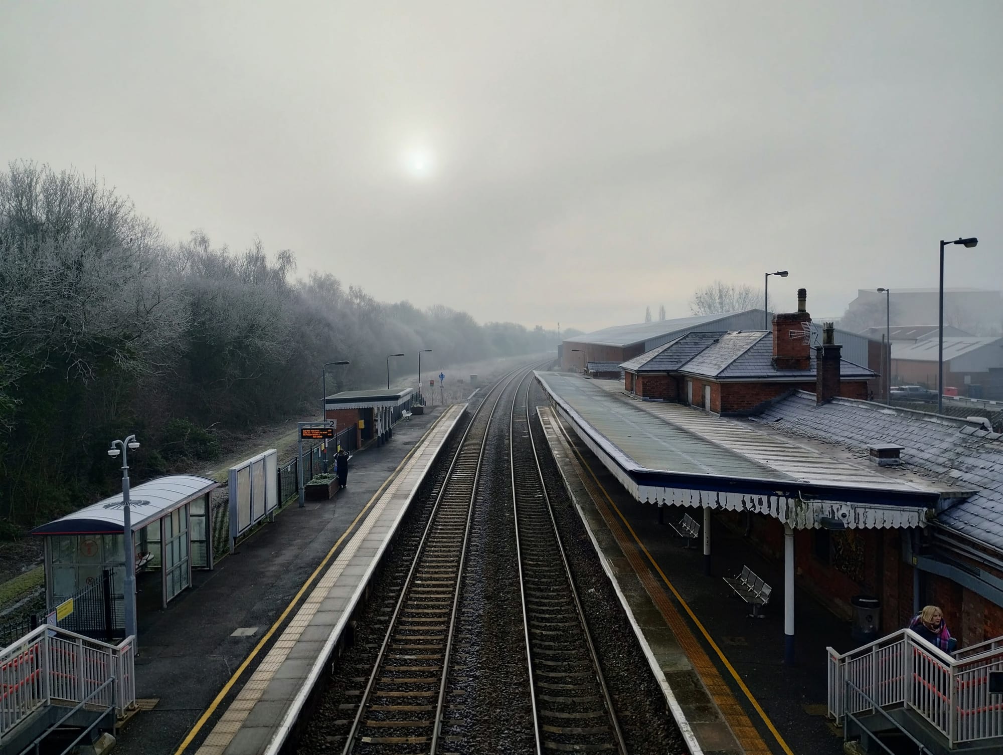 Looking down the tracks from the a footbridge, frosted trees and roofs either side of a wintry railway station.