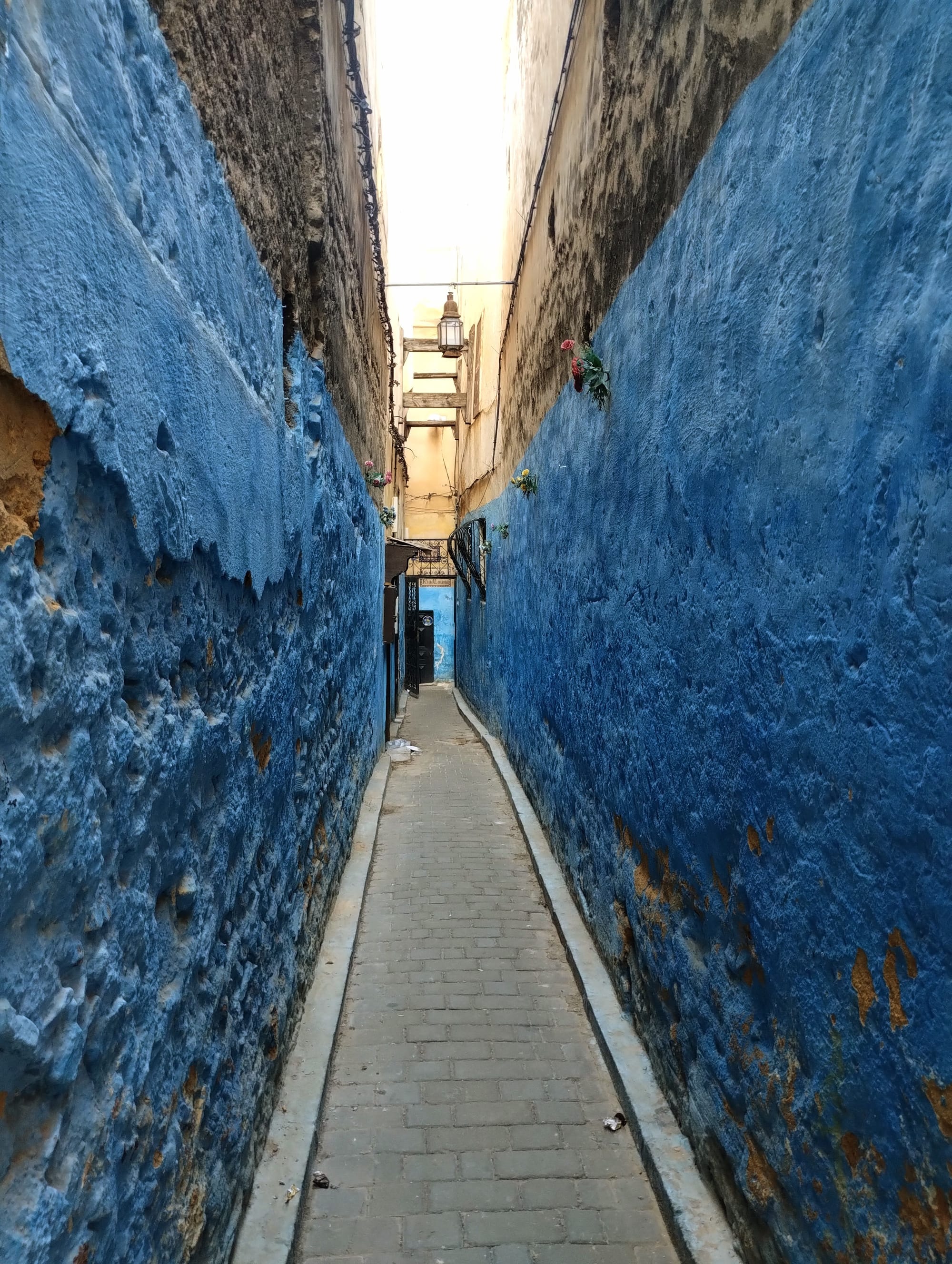 Narrow alleyway with vibrant blue walls and a brick path, leading towards a bright opening in the distance.