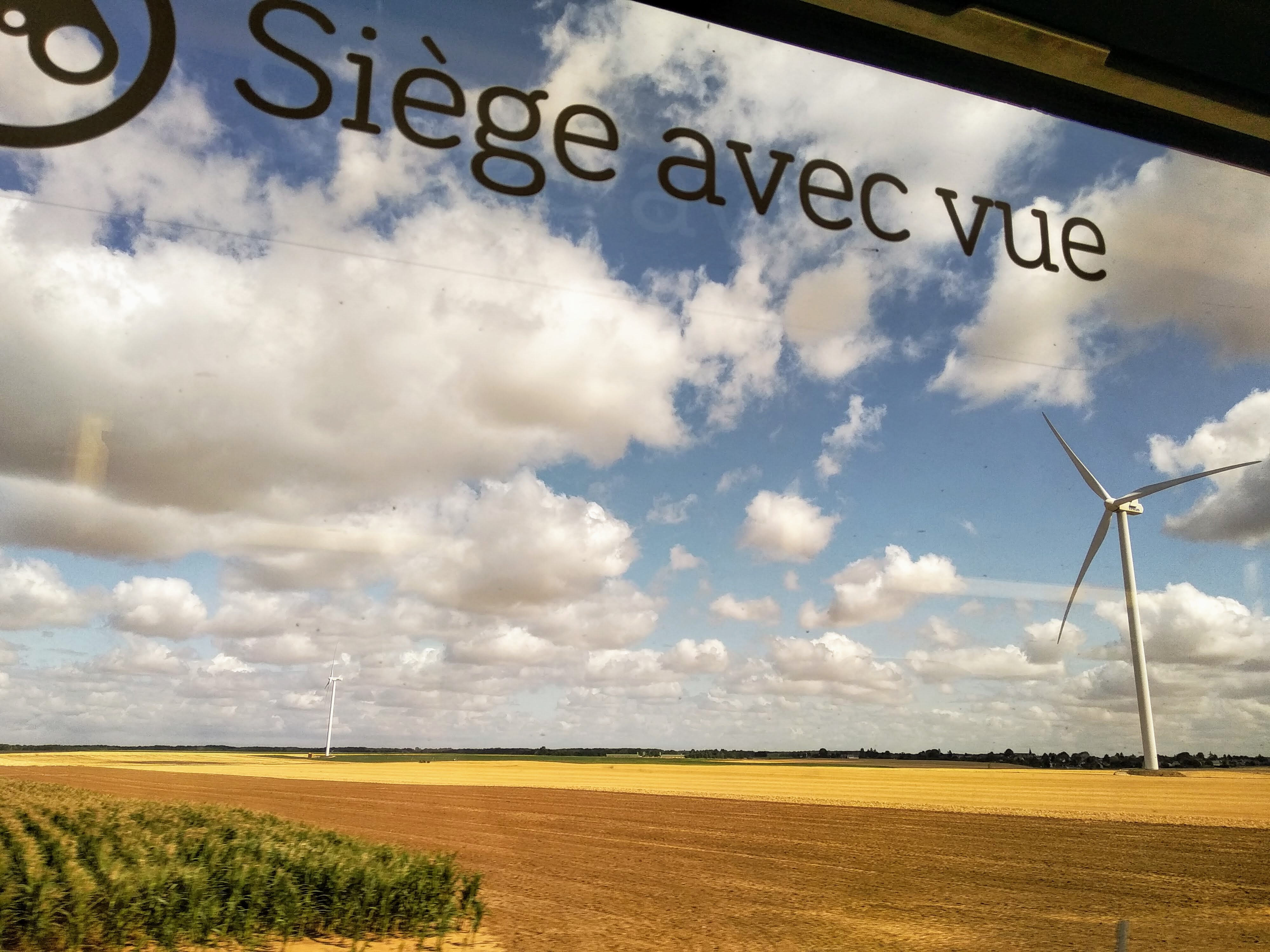 View from a train window of a blue sky filled with white puffy clouds and wind turbines in a field of straw, with the French phrase "Siège avec vue" visible on the window's surface.