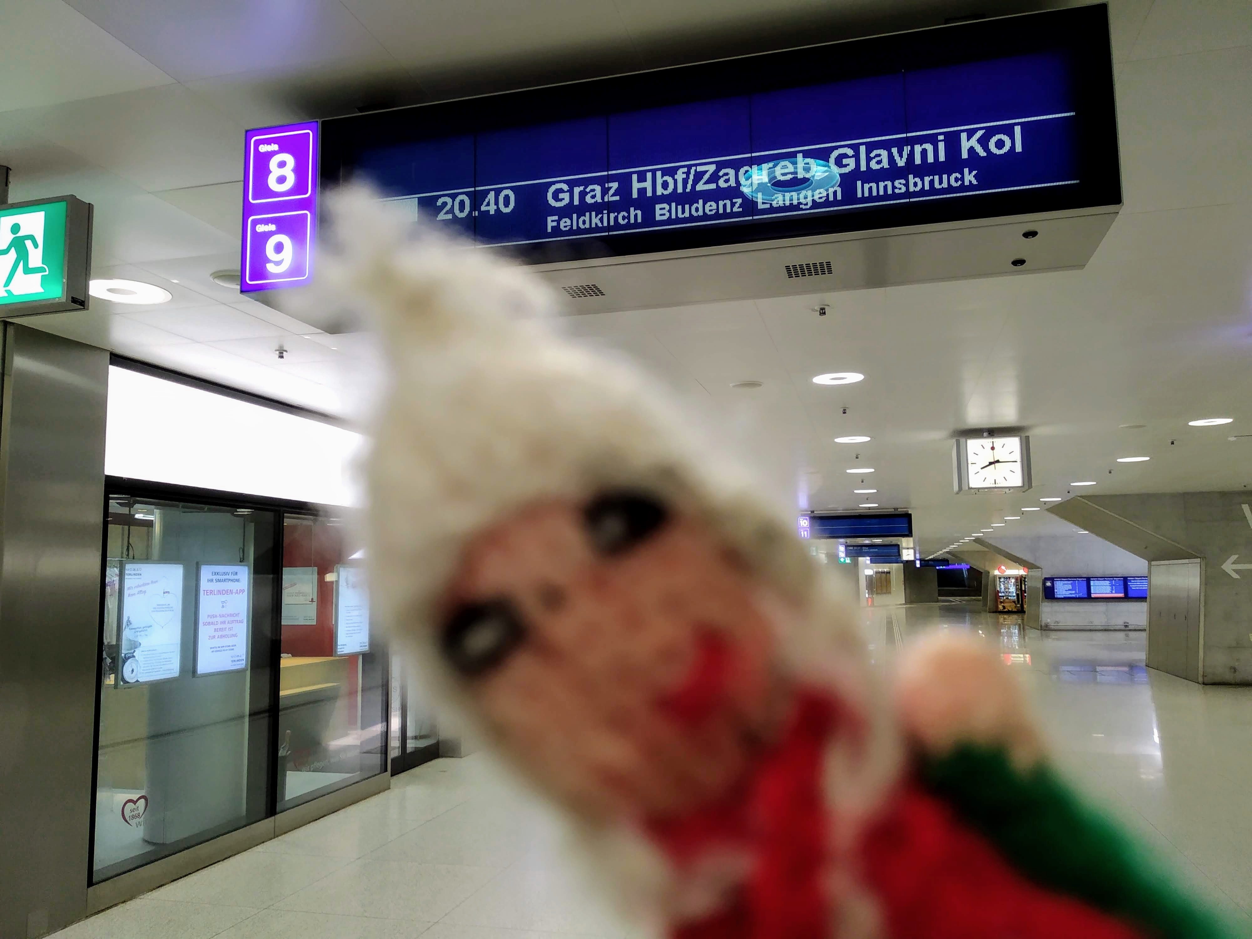 A blurry finger puppet in an empty Zürich HBF. An illuminated departure board lists a night train at 20:40 to Graz Hbf/Zagreb from platform 9. In the background, a clock reads 20:15.