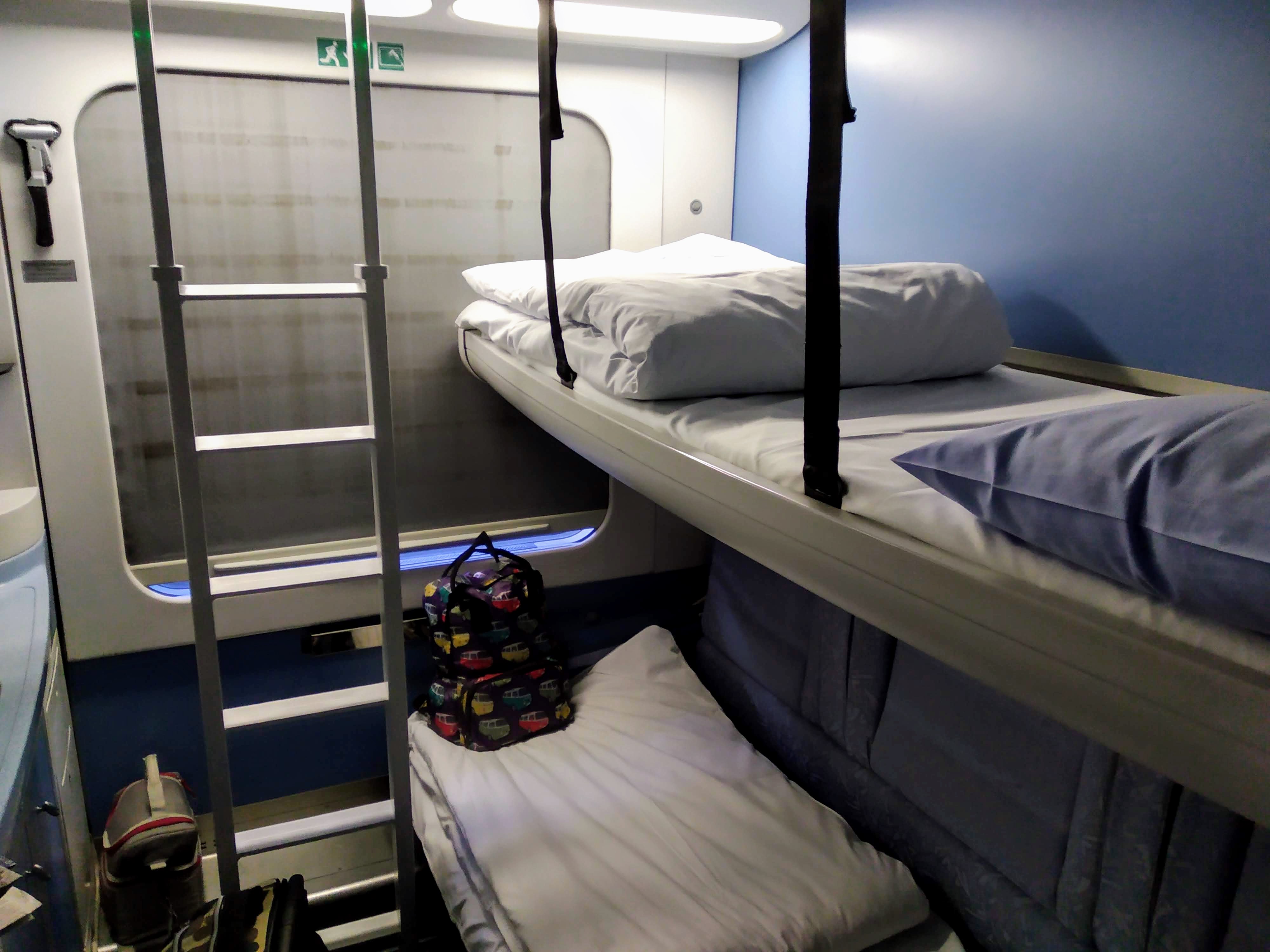 A bunk bed in a train sleeper car, the upper berth has a white blanket and pillow, the lower berth has a white blanket and a blue pillow. A ladder allows access the upper berth. There is a bag on the lower berth, beneath the window. The curtain is closed.