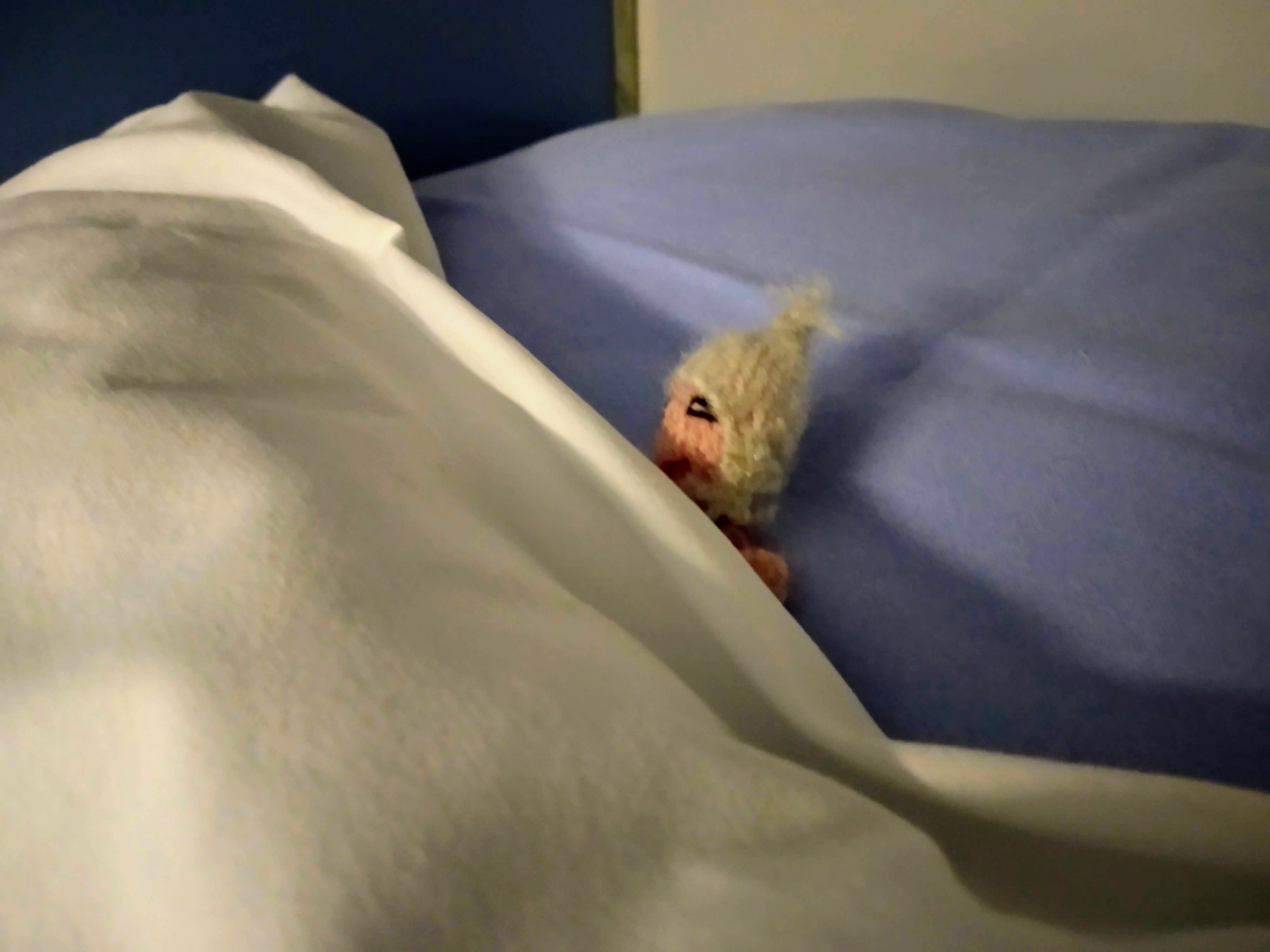 A finger puppet tucked up in bed against a blue pillow, under a white duvet.