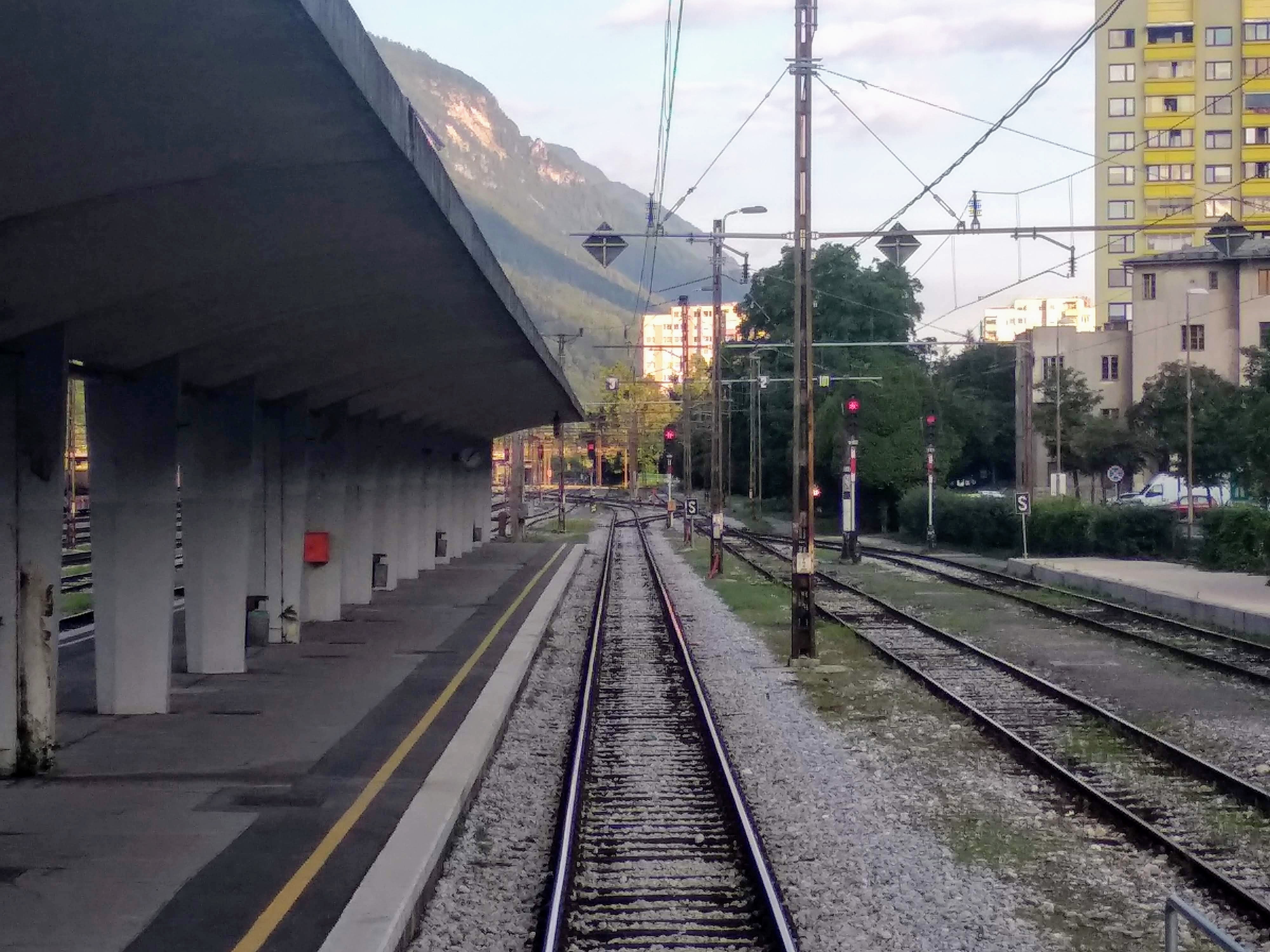 View down a railway platform with multiple tracks; a concrete canopy is on the left, supported by pillars; tracks lead into the distance past red signal lights; a mountain is visible in the background. Residential buildings on the right.