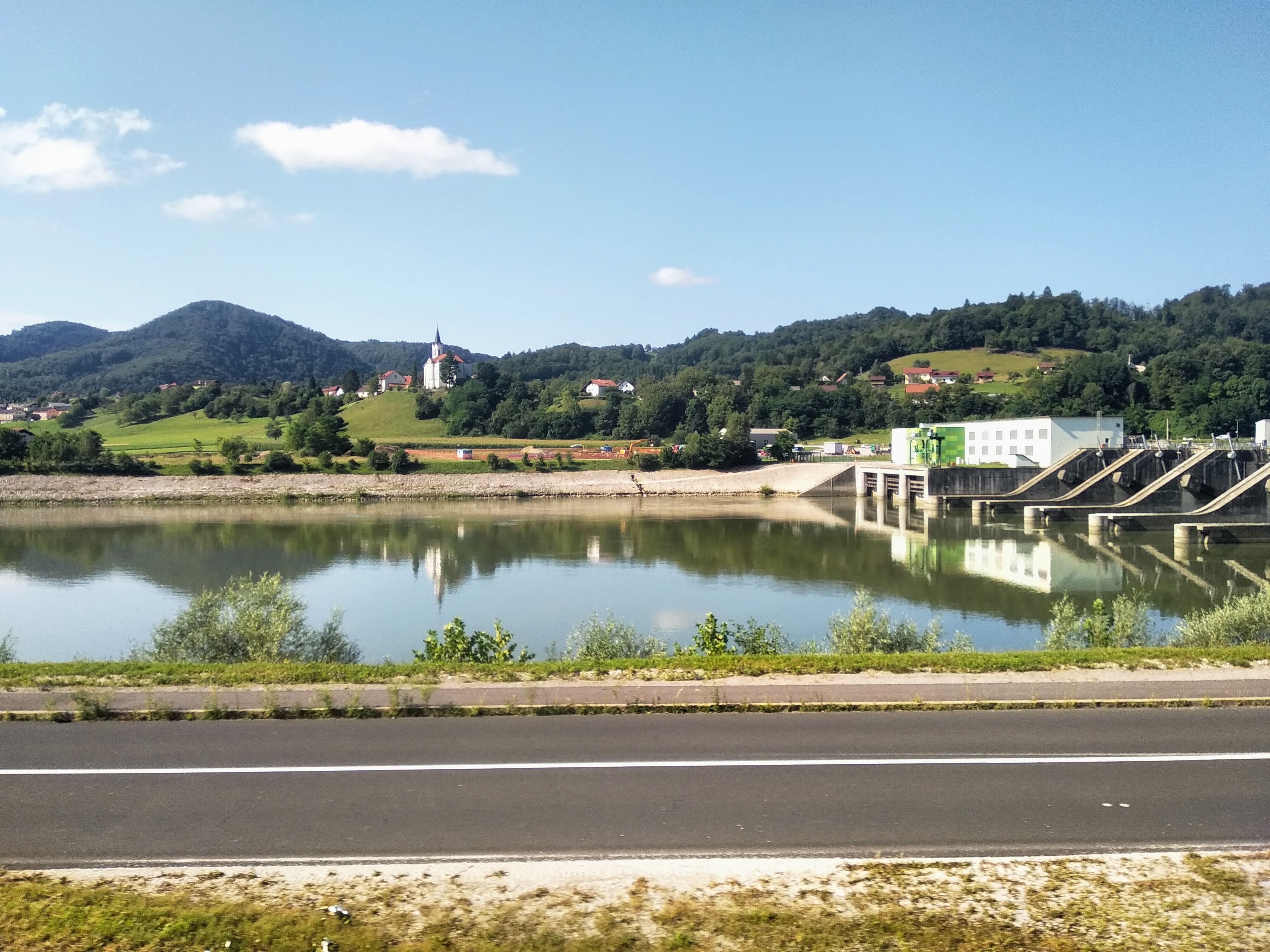 Wide shot of a lake with a dam, green hills, and a church steeple in the background, under a blue sky.