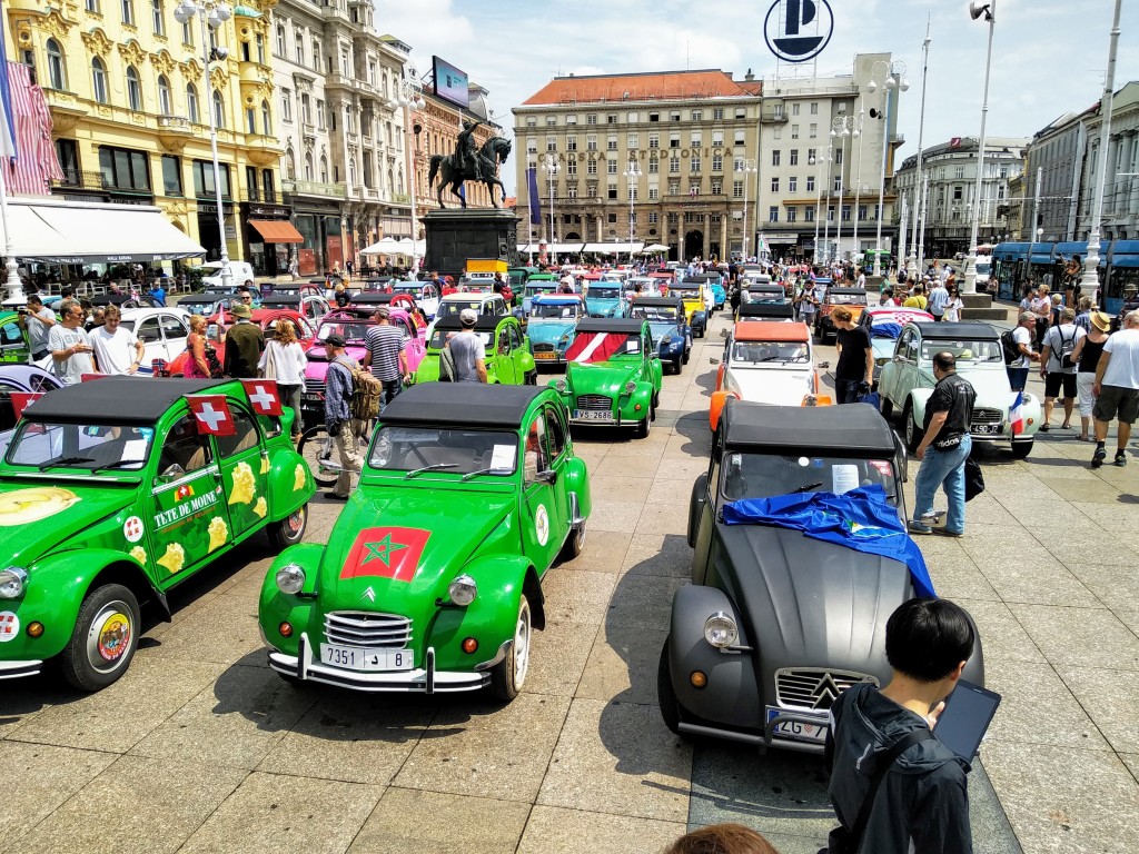 A large gathering of vintage Citroën 2CV cars in a town square, many brightly coloured with various flags and graphics. People milling about and historic buildings in the background. A prominent statue of a man on a horse stands behind the cars.