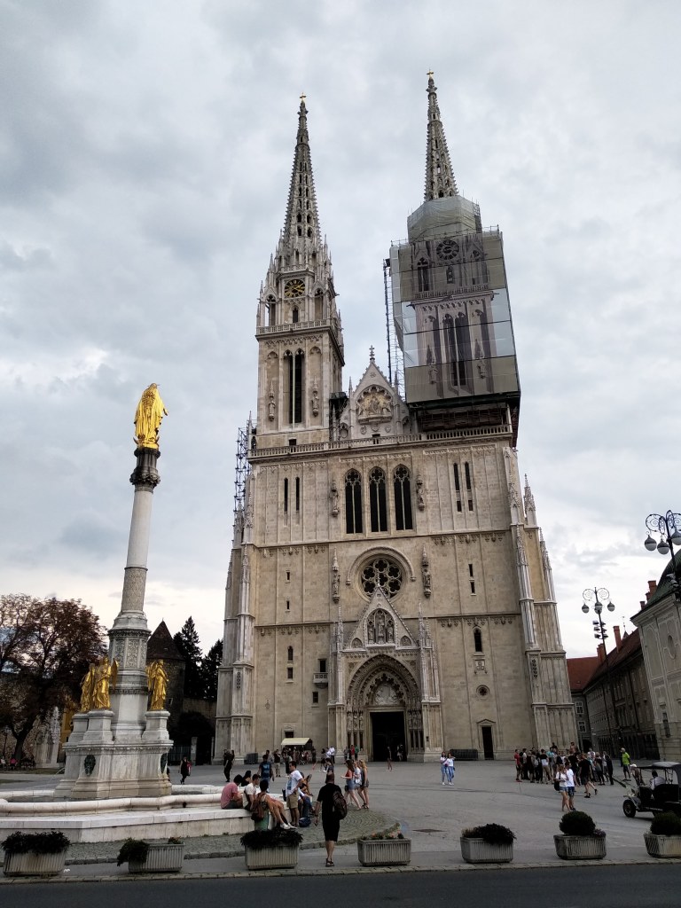 A low-angle shot of the cathedral in Zagreb, a gothic style church with two tall spires under a cloudy sky. A gilded Marian column is positioned on the left. Many tourists populate the square. One spire is covered with scaffolding.