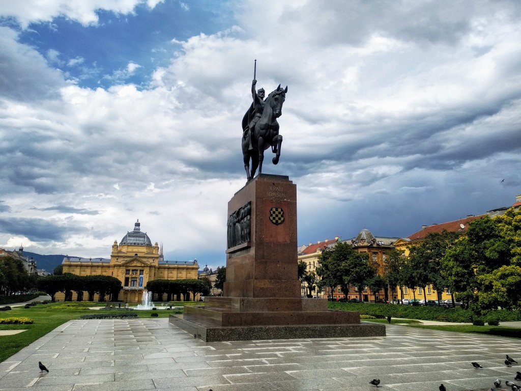 Statue of King Tomislav on horseback in Zagreb, Croatia, atop a large pedestal in a public square, with the Arts Pavilion and other buildings in the background under a dramatic cloudy sky.