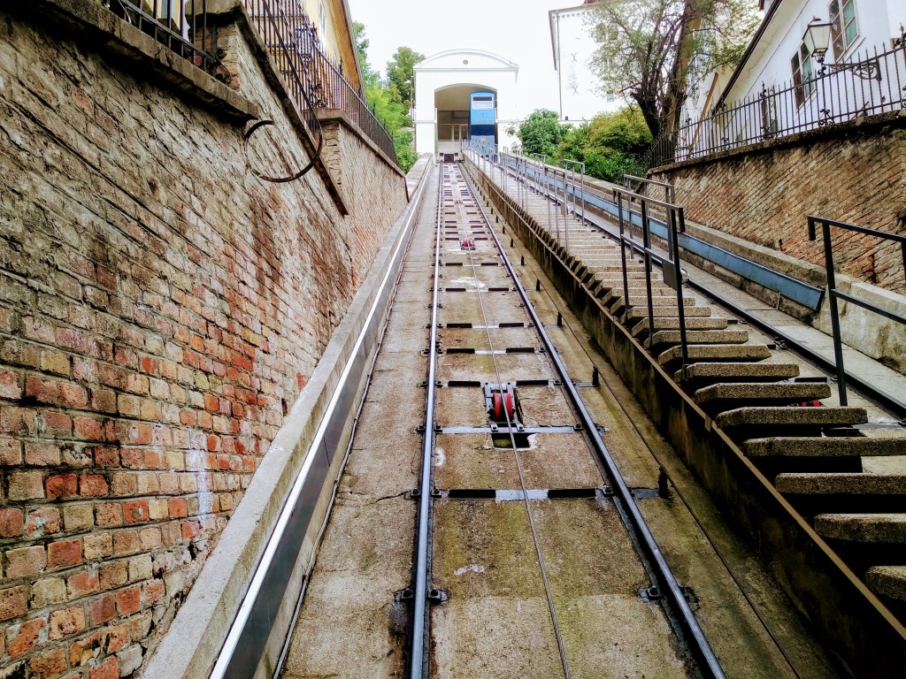 An ascending perspective of a funicular railway track leading uphill to a station, flanked by brick walls and a staircase. A control mechanism is visible along the track. At the top of the track is a station building.