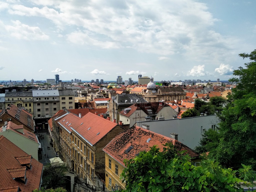 Overhead view of Zagreb, Croatia, featuring red-tiled rooftops, various architectural styles, a bustling cityscape, and a cloudy sky.