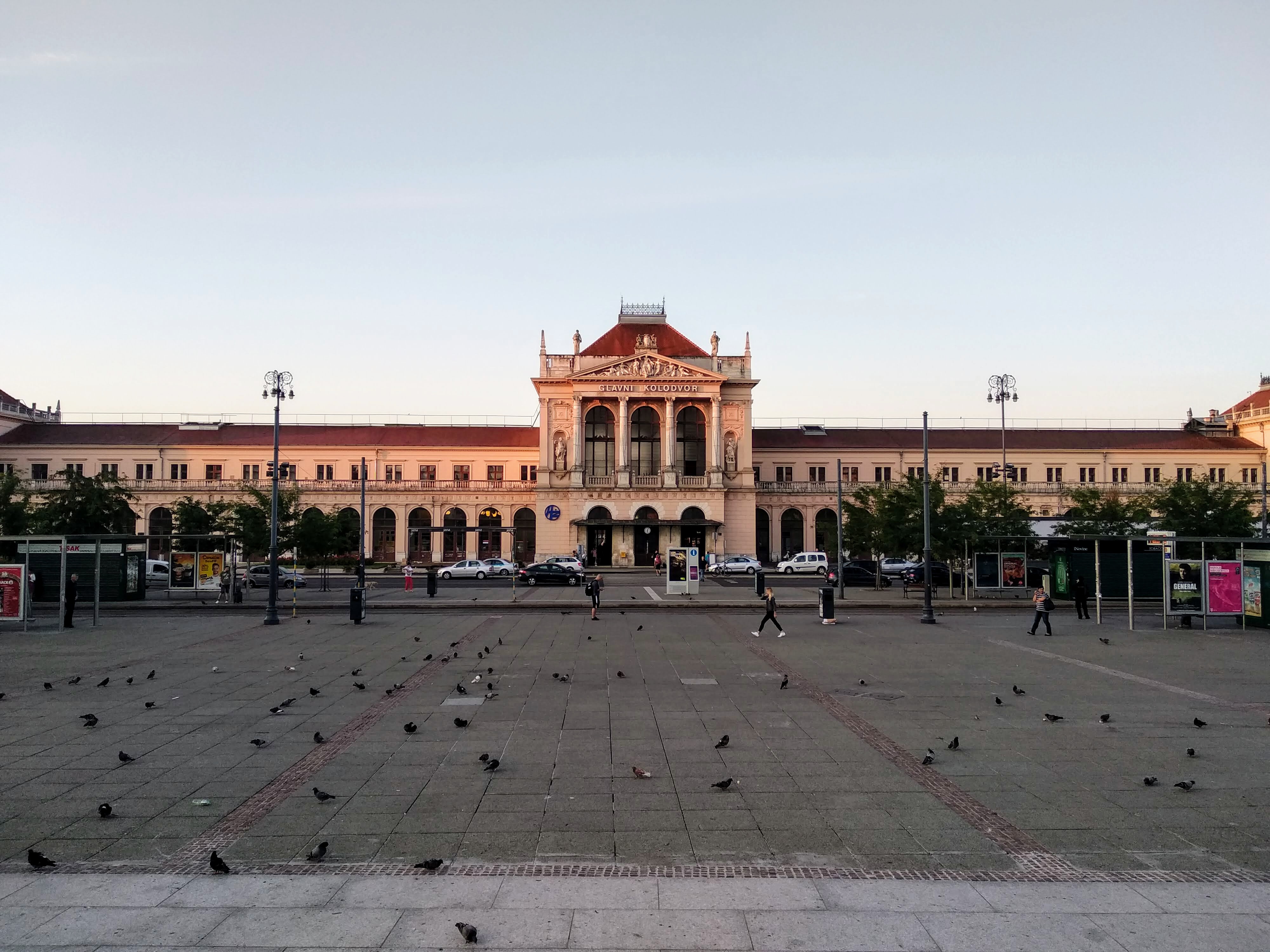 Exterior view of sunlit Zagreb Central Station under a morning sky, with pedestrians, vehicles, and a squadron of pigeons on the plaza in front of the building.
