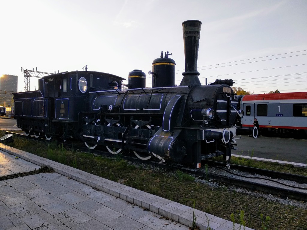 A black vintage steam locomotive on tracks at a station with a red and white train car in the background.
