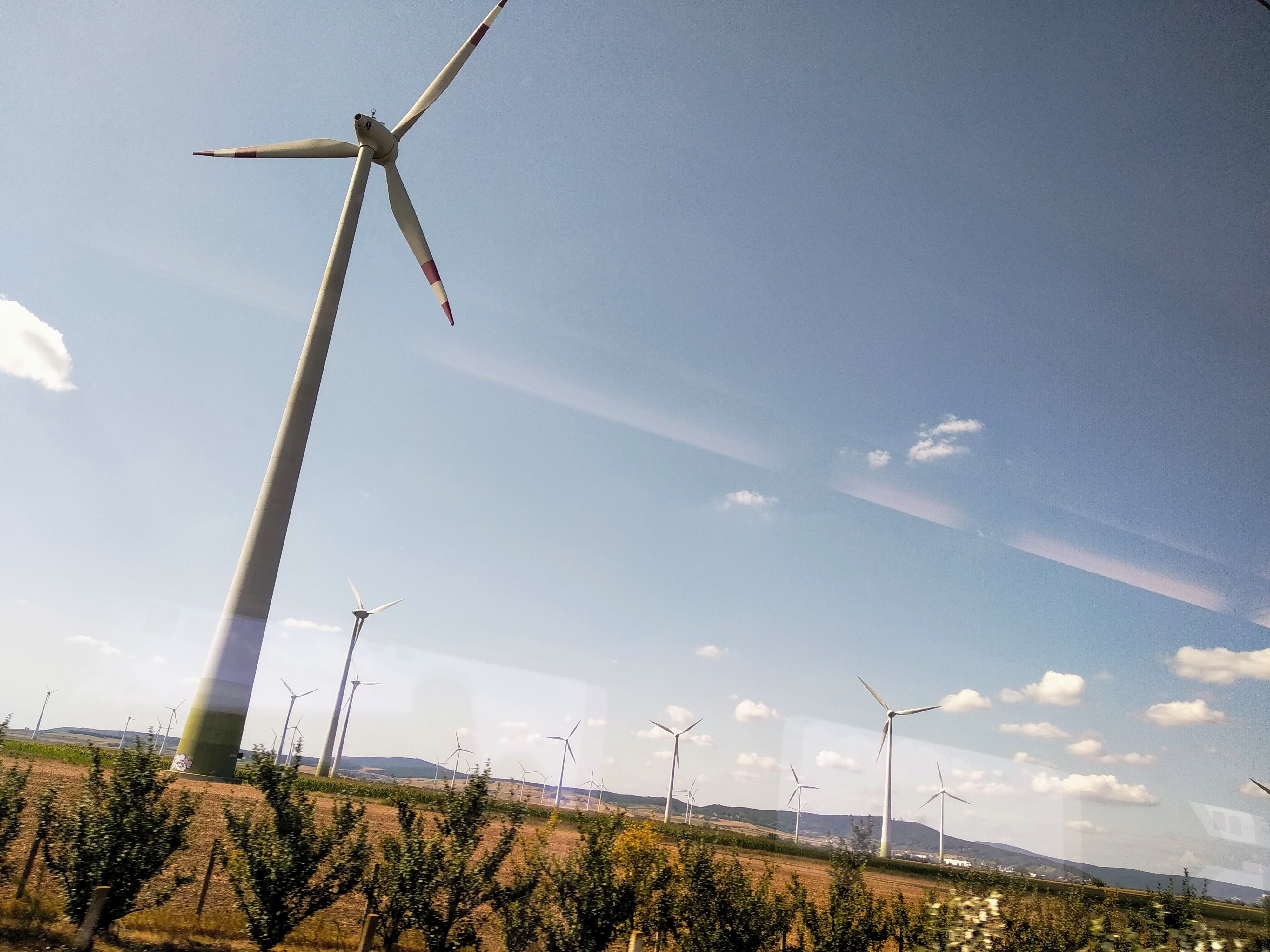 A field of wind turbines under a partly cloudy, blue sky. The tall, white turbines with red-tipped blades stand in a field of crops and trees, with the distant hills in the background. The image is framed from inside a train, through a slightly reflective window.