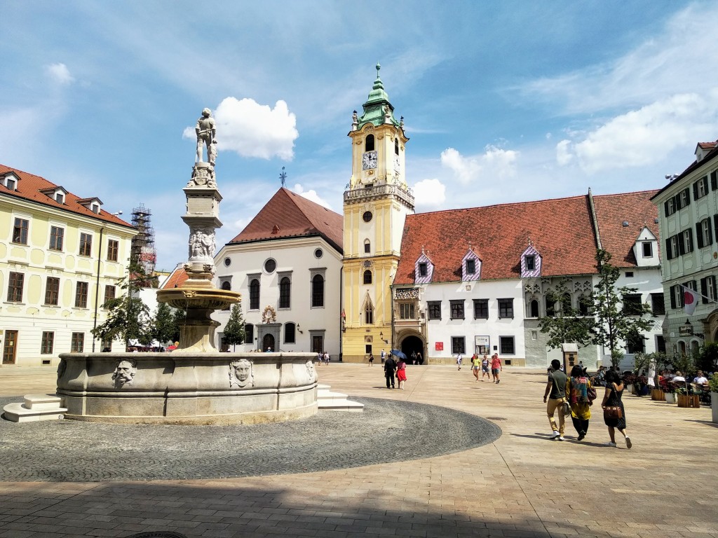 A sunny day in a square with a baroque fountain as the focal point, featuring a statue atop a tall column. Behind the fountain are historic buildings including a clock tower and a building with a red-tiled roof. People are walking around the square, some under parasols. The sky is blue with fluffy white clouds.