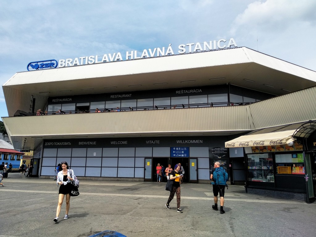 Exterior view of Bratislava Hlavná Stanica train station under a partly cloudy sky, with multilingual signage indicating welcome and restaurants above the entrance. Several people walk around the plaza in front.