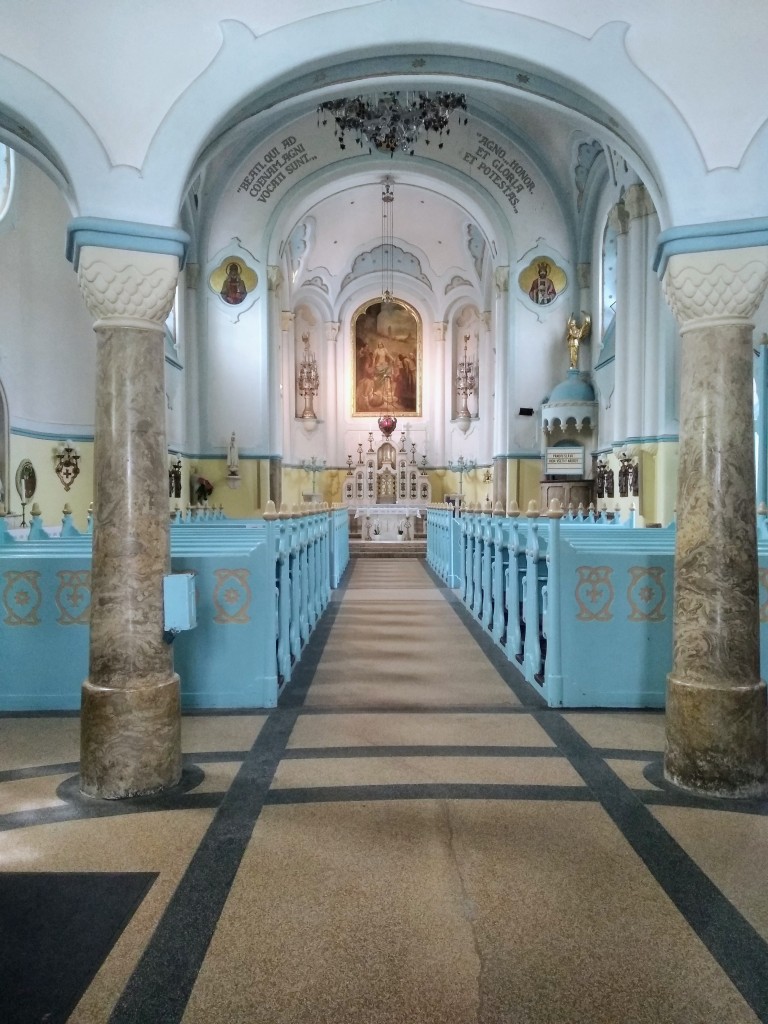 Interior view of a church with an aisle lined by light blue pews and ornate marble columns.  The perspective leads to a raised altar under a painted mural, illuminated by natural light, and a decorative ceiling.