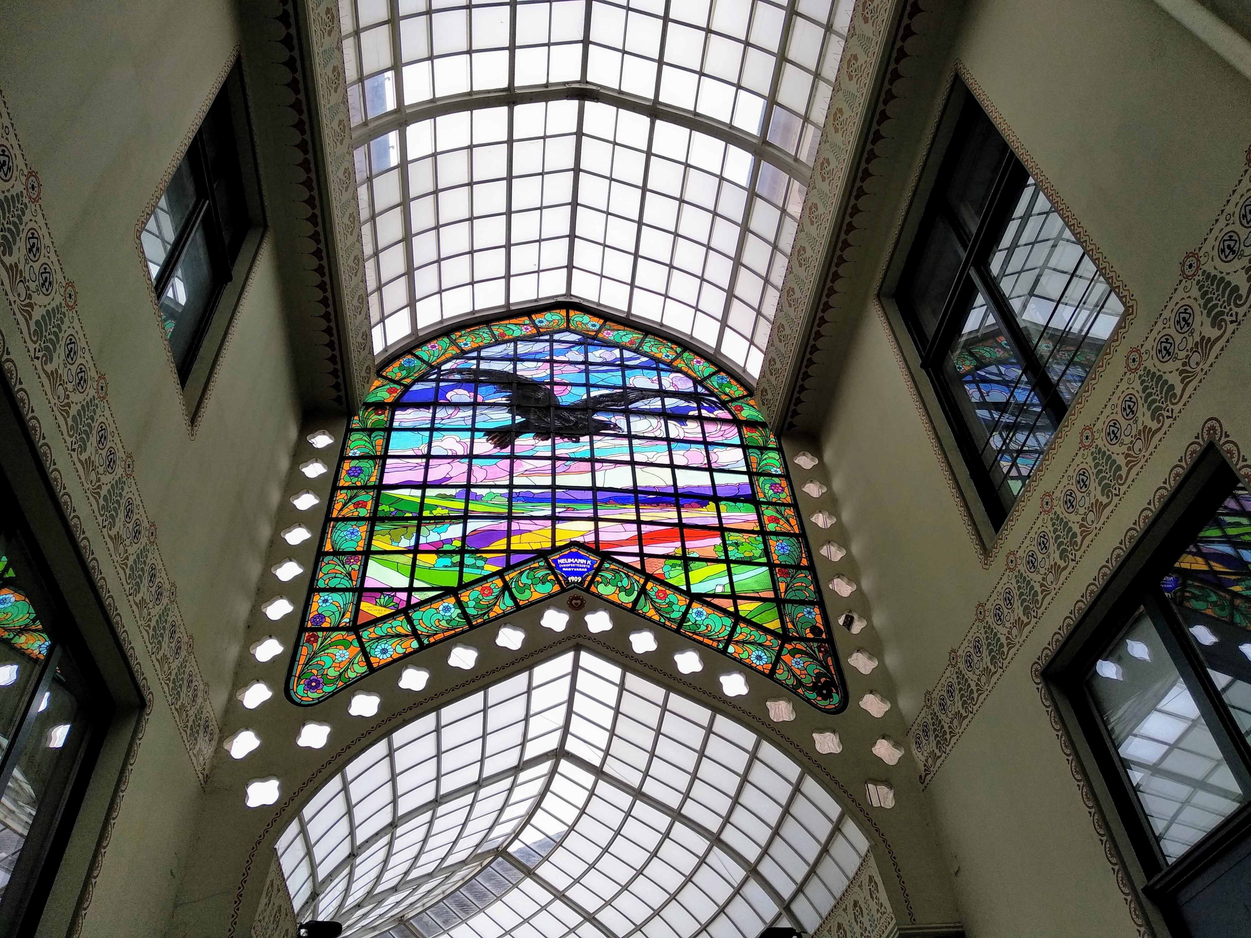 Interior view looking up at a stained-glass window depicting a black bird in flight against a colourful, abstracted landscape background, arched over a grid-patterned skylight, flanked by decorative, windowed walls and a repetitive floral trim.