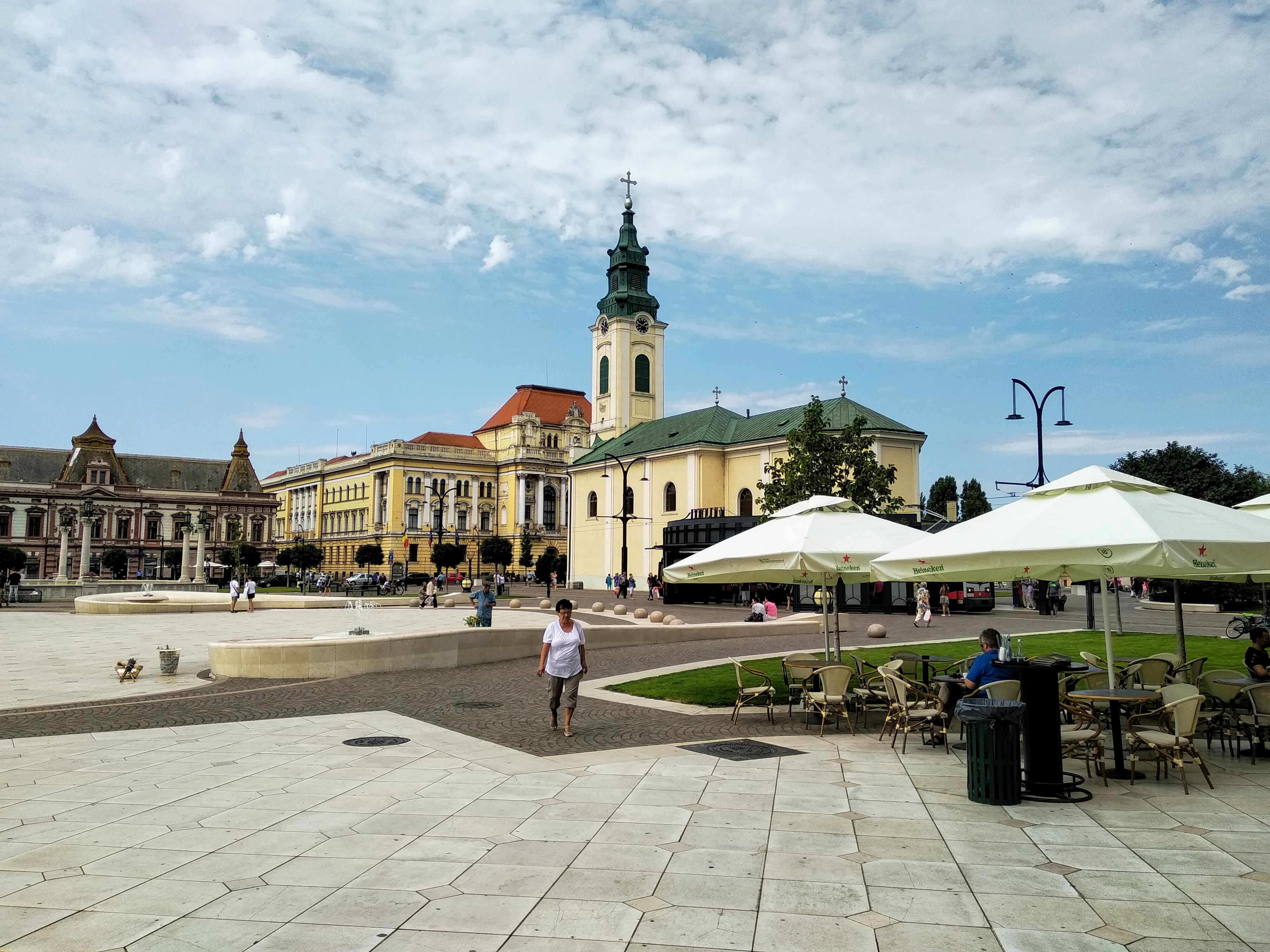 View of a sunlit European town square dominated by a yellow and green church with a tall clock tower, outdoor cafe with umbrellas, and several ornate buildings under a blue sky with little fluffy clouds.