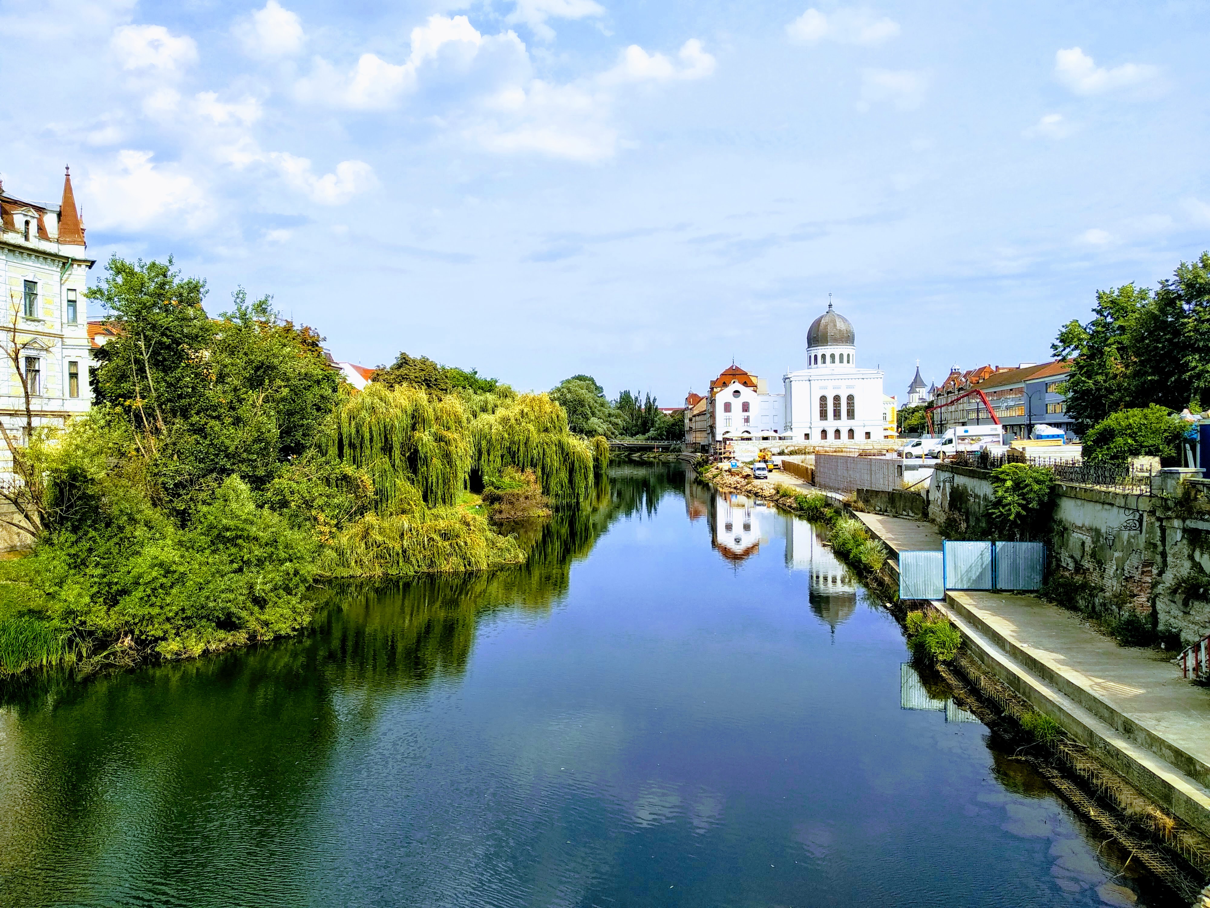 Scenic view of a wide, calm river reflecting the blue sky and puffy clouds, flanked by lush green trees and historic buildings, including a white domed synagogue.