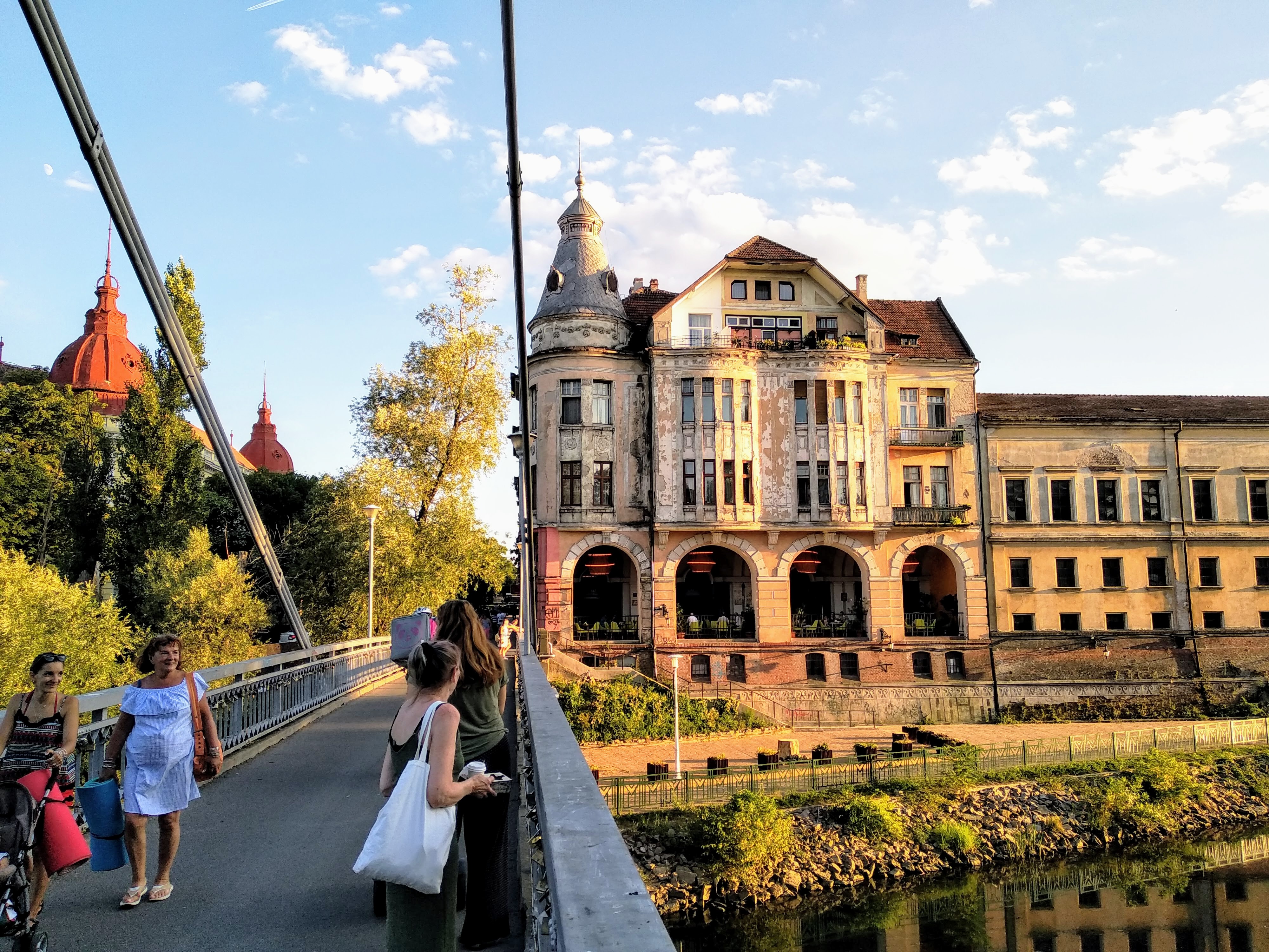 People walk along a suspension bridge in front of a historic building with an arched facade and a domed tower, with red-domed buildings visible in the background, and a river below.