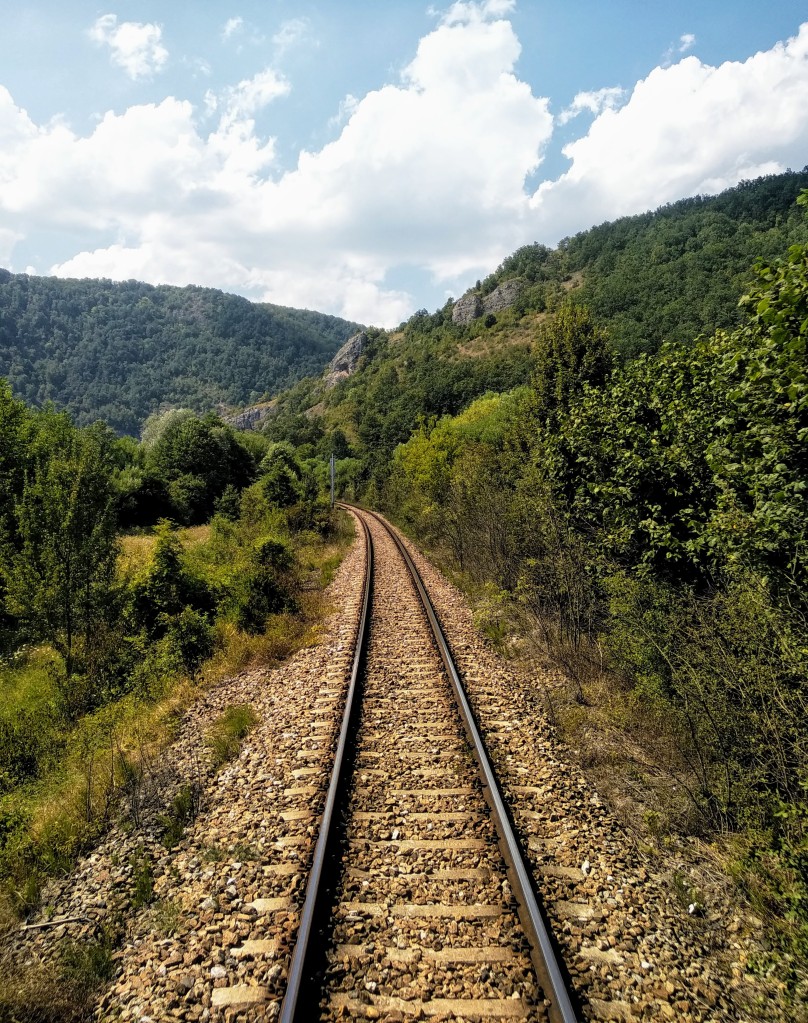 Curving railway tracks through a lush green valley under a blue sky with puffy white clouds, surrounded by dense vegetation and hills.