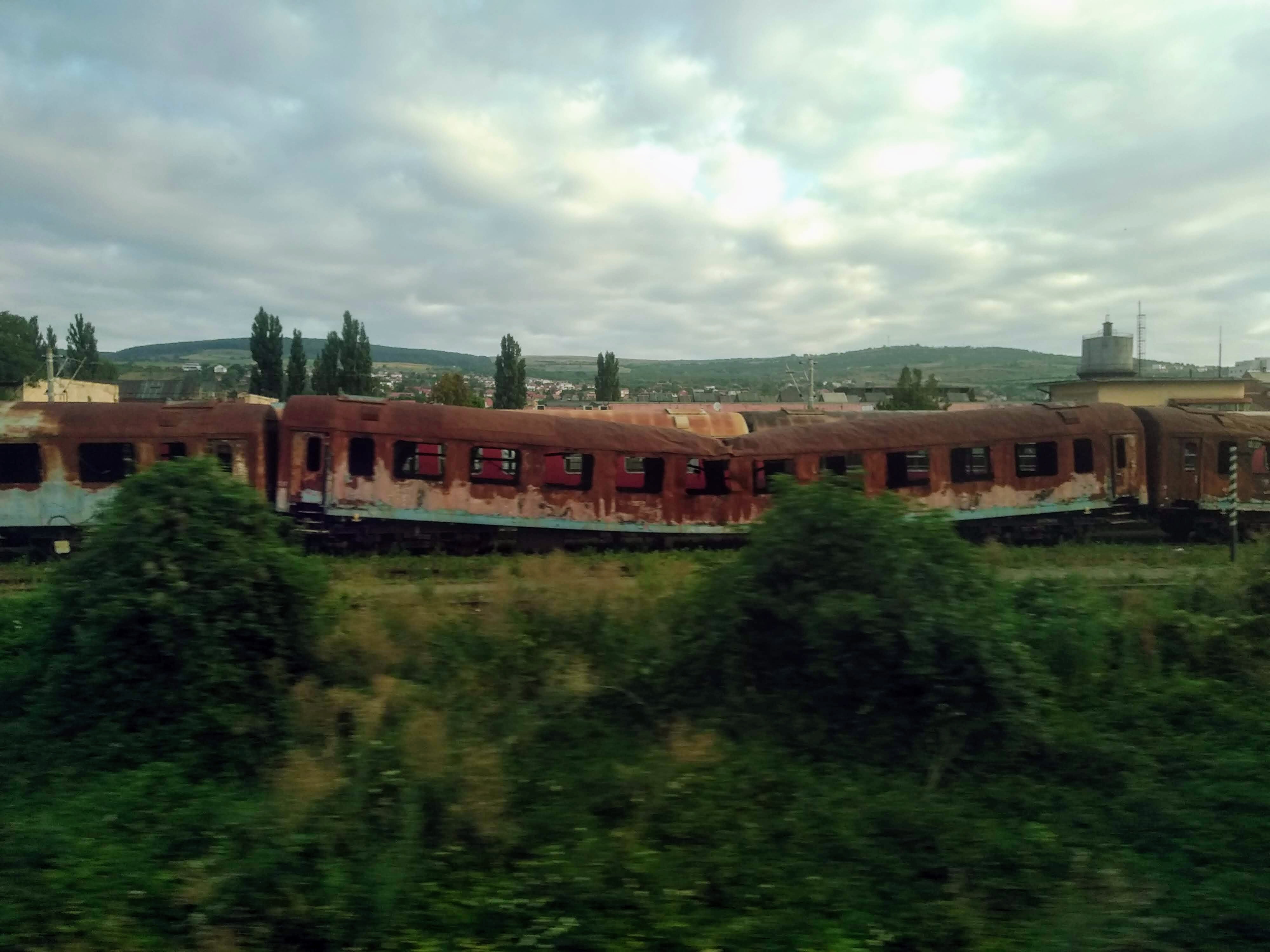 Rusty, abandoned train carriages sit on tracks surrounded by overgrown vegetation against a cloudy sky with a distant town in the background.