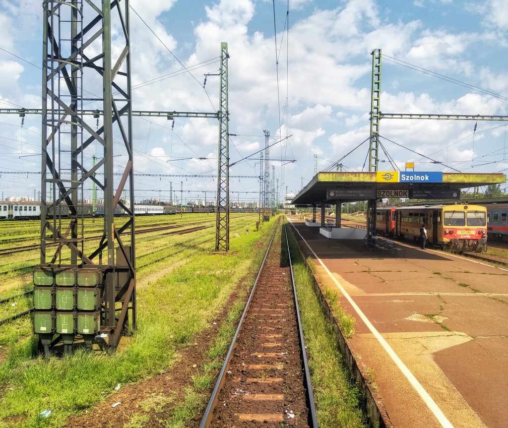 Szolnok railway station in Hungary, with train tracks in the foreground leading to a station platform with a yellow and red train. Overhead power lines and a sign with the station's name are visible. Other train cars are present in the background alongside parallel tracks amidst grassy areas.