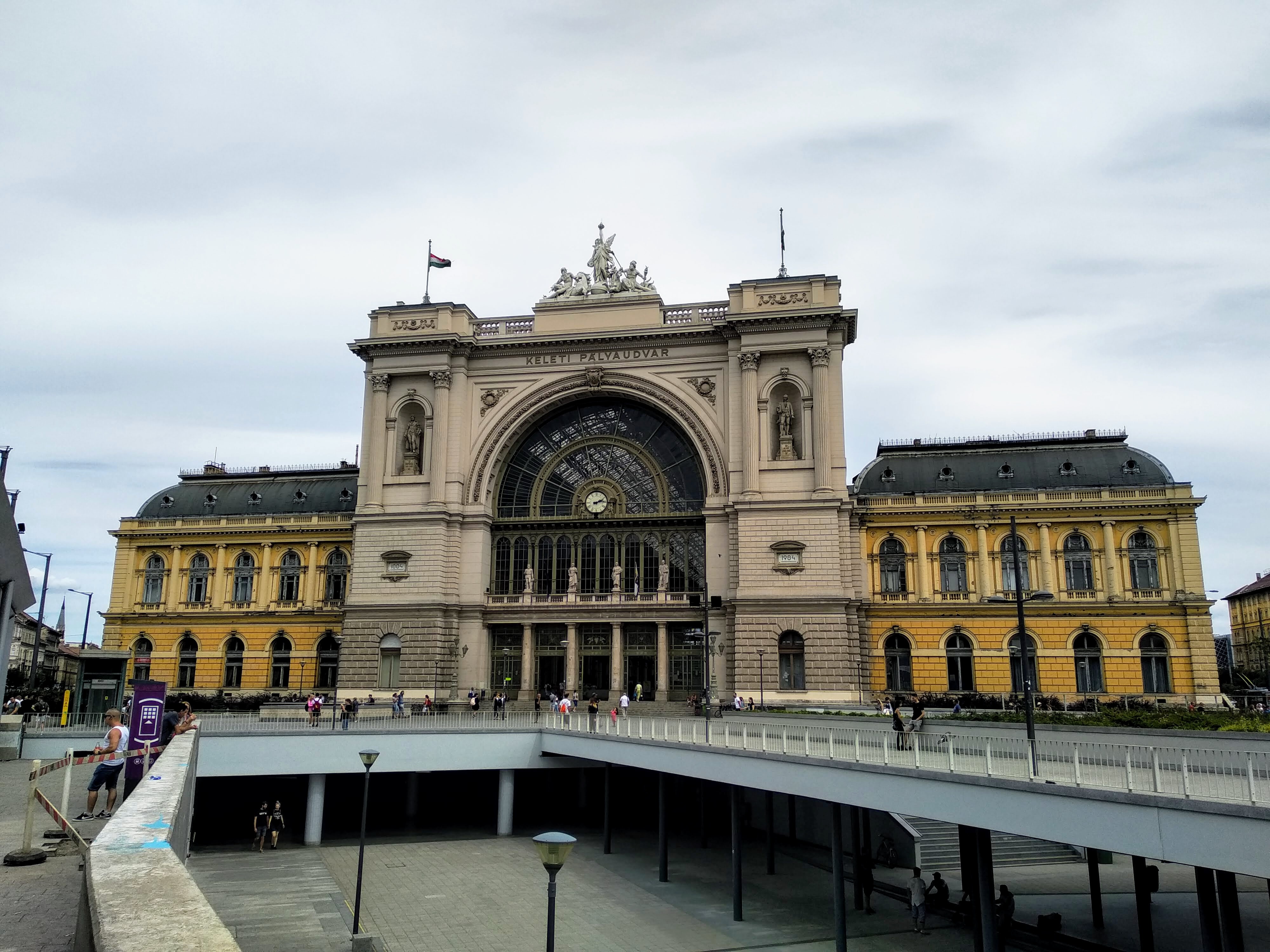 Exterior view of Keleti Pályaudvar (Eastern Railway Station) in Budapest, Hungary, featuring a large, beige neoclassical facade with arched windows, a prominent glass and steel arched entrance, a clock, sculptures, and numerous small flags. Pedestrians walk across a bridge in front of the station.