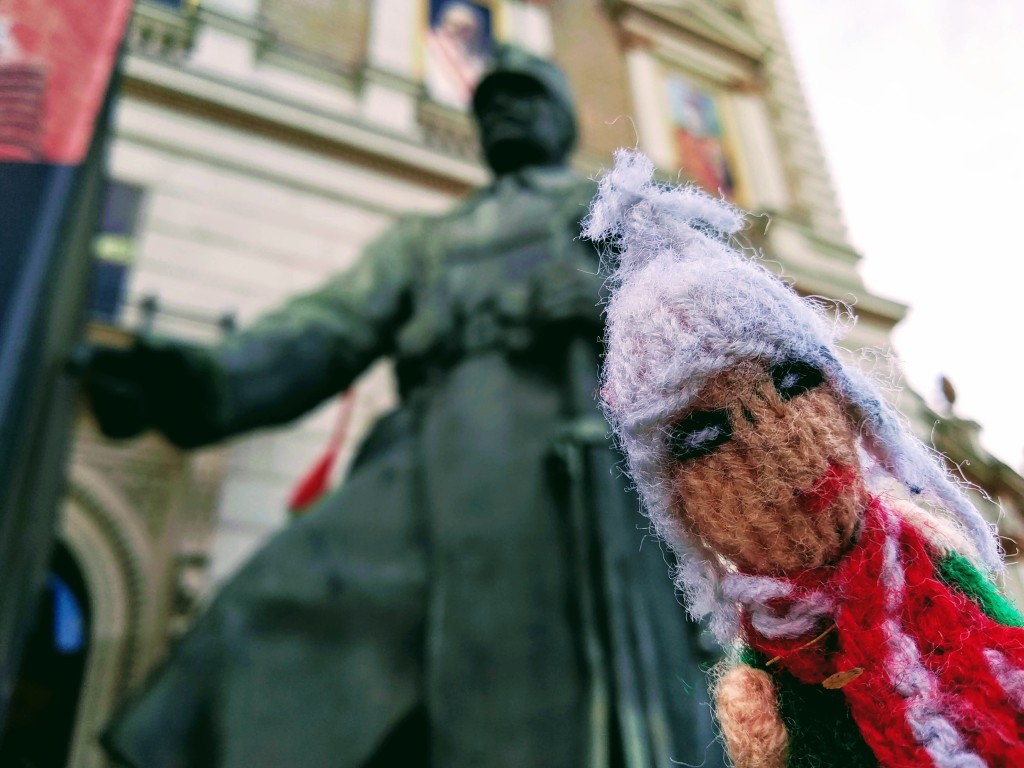 Close-up shot of a weary finger puppet in front of a blurred bronze statue of a soldier in a long coat.