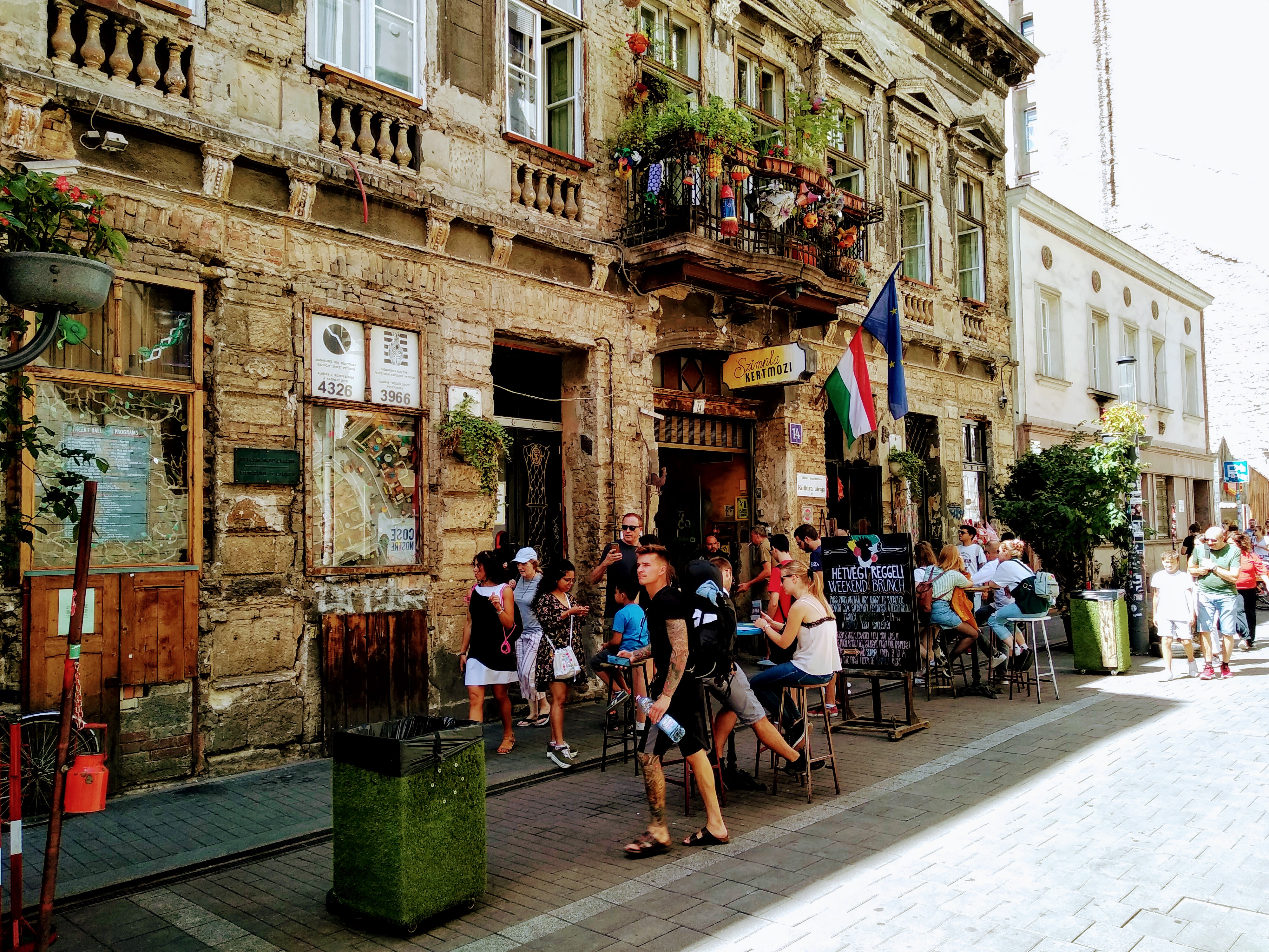 Exterior view of a historic building in Budapest with a pavement cafe area. A narrow brick building with a balcony adorned with plants and a sign for "Szimpla Kertmozi" is shown. People are standing, walking, and sitting at tables on the sidewalk in front of the cafe. Flags of Hungary and the European Union hang near the entrance.