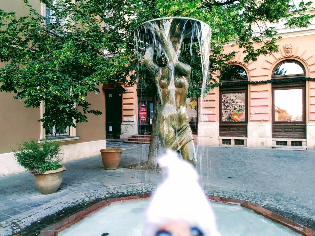 Bronze nude woman sculpted in the cold with water cascading from a bowl held above her head, set in a cobblestone plaza with buildings and greenery in the background. A finger puppet looks aroused.