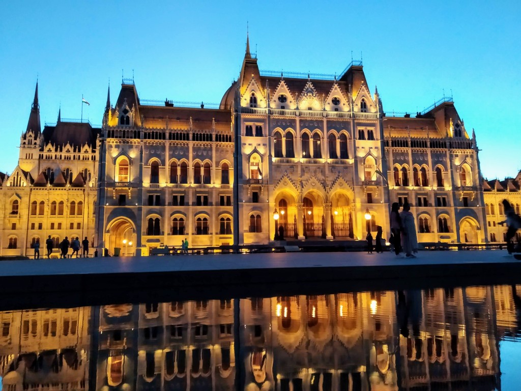 The illuminated Hungarian Parliament Building at dusk, reflected in the calm waters of the Danube River, with silhouetted figures of people on the embankment.