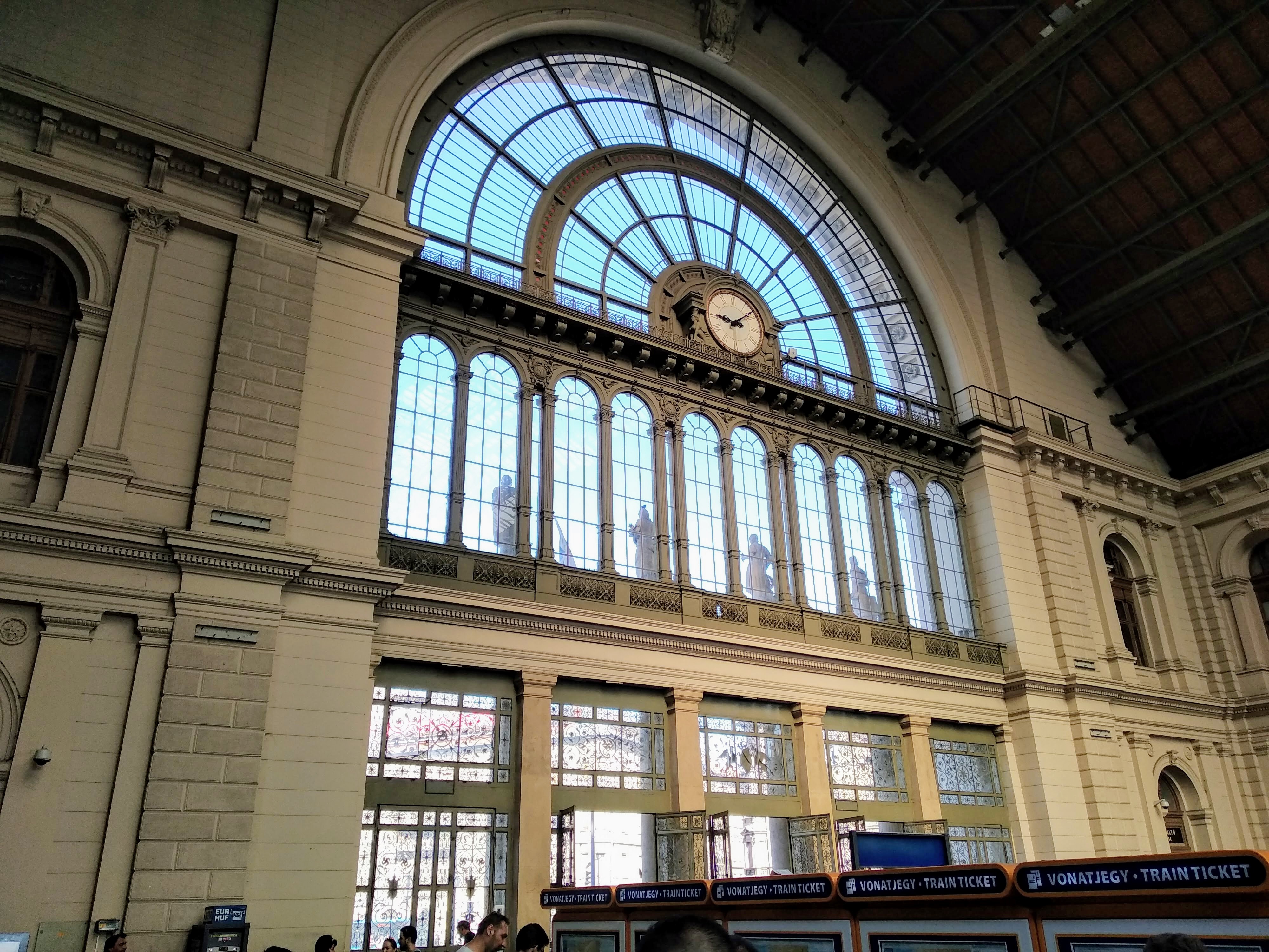 Interior view of a grand train station featuring a large arched window with a clock at the centre, smaller arched windows below with statues, and five orange ticket machines.