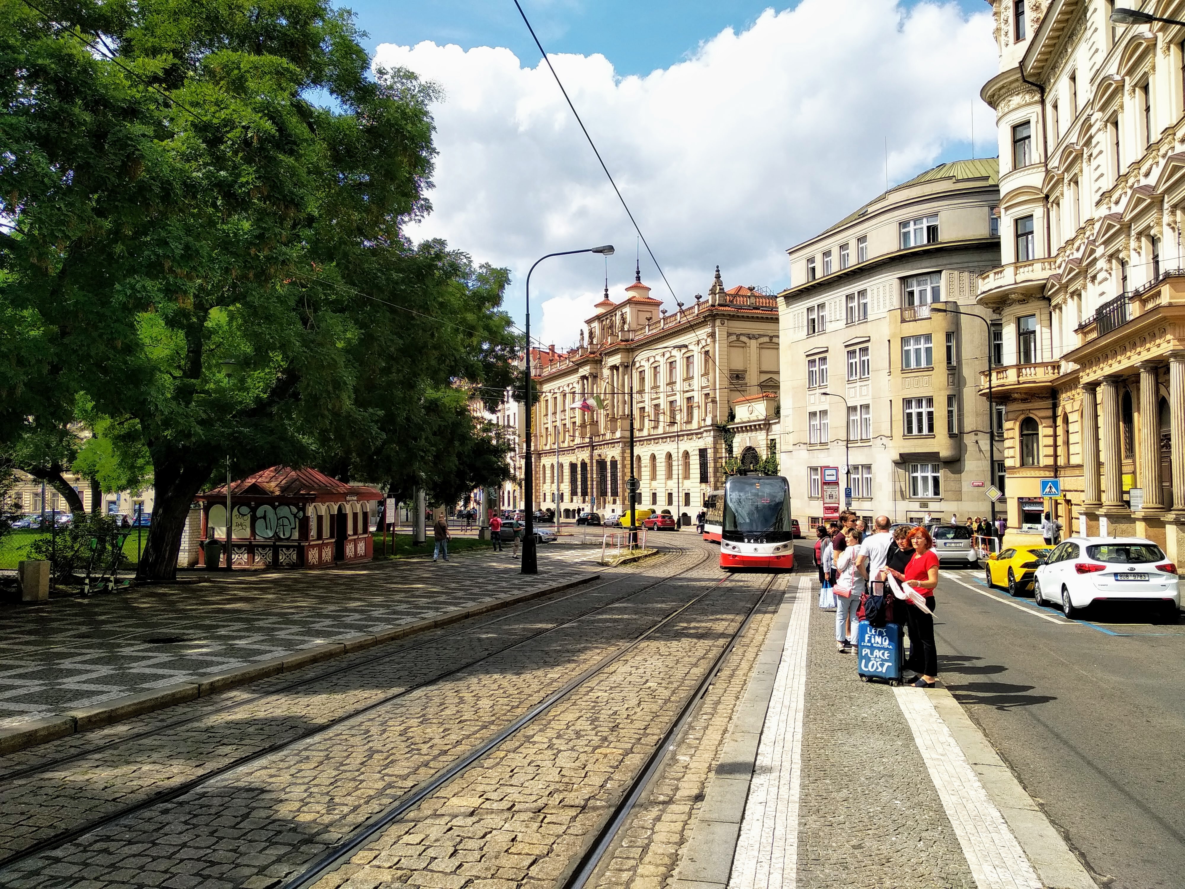 Street scene with tram tracks, a red and white tram, people waiting at a stop, and grand European architecture under a sunny sky.