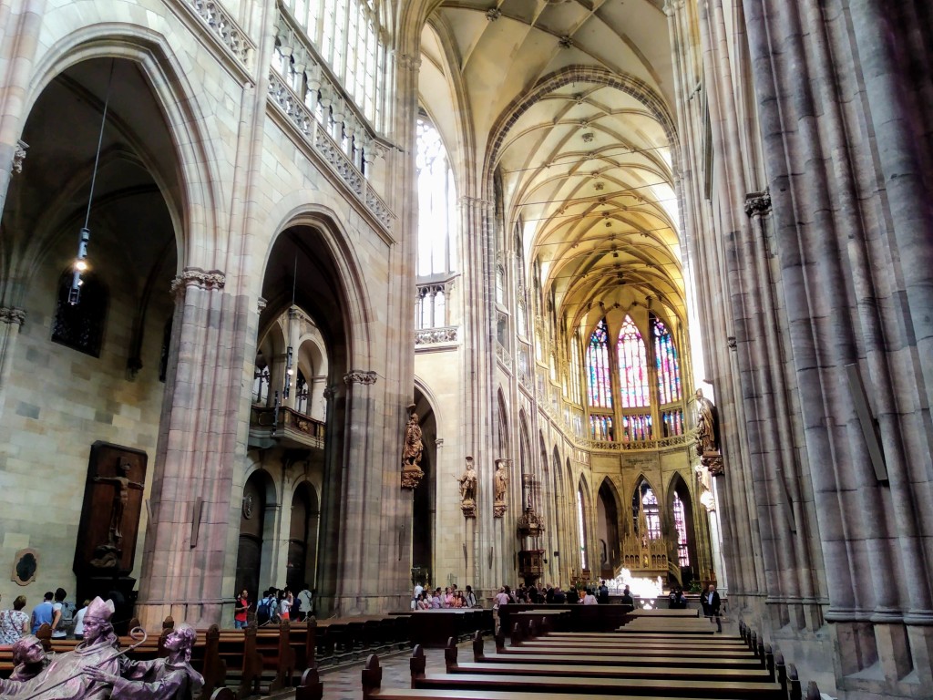 Interior of a large cathedral looking towards the chancel, with a high vaulted ceiling, massive stone pillars, arched doorways, and rows of wooden pews leading towards a stained-glass windowed apse.