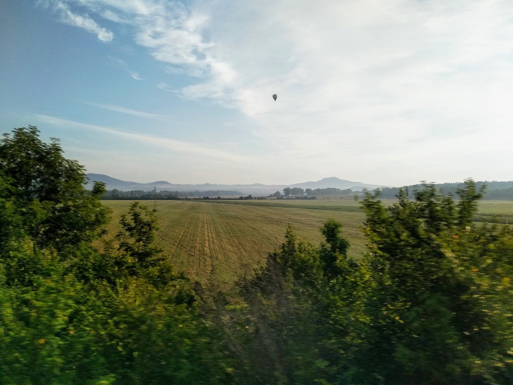 A scenic view of a harvested agricultural field crossed with tractor tracks, surrounded by greenery, with mountains in the distance under a partly cloudy blue sky. A solitary hot air balloon floats in the upper centre.