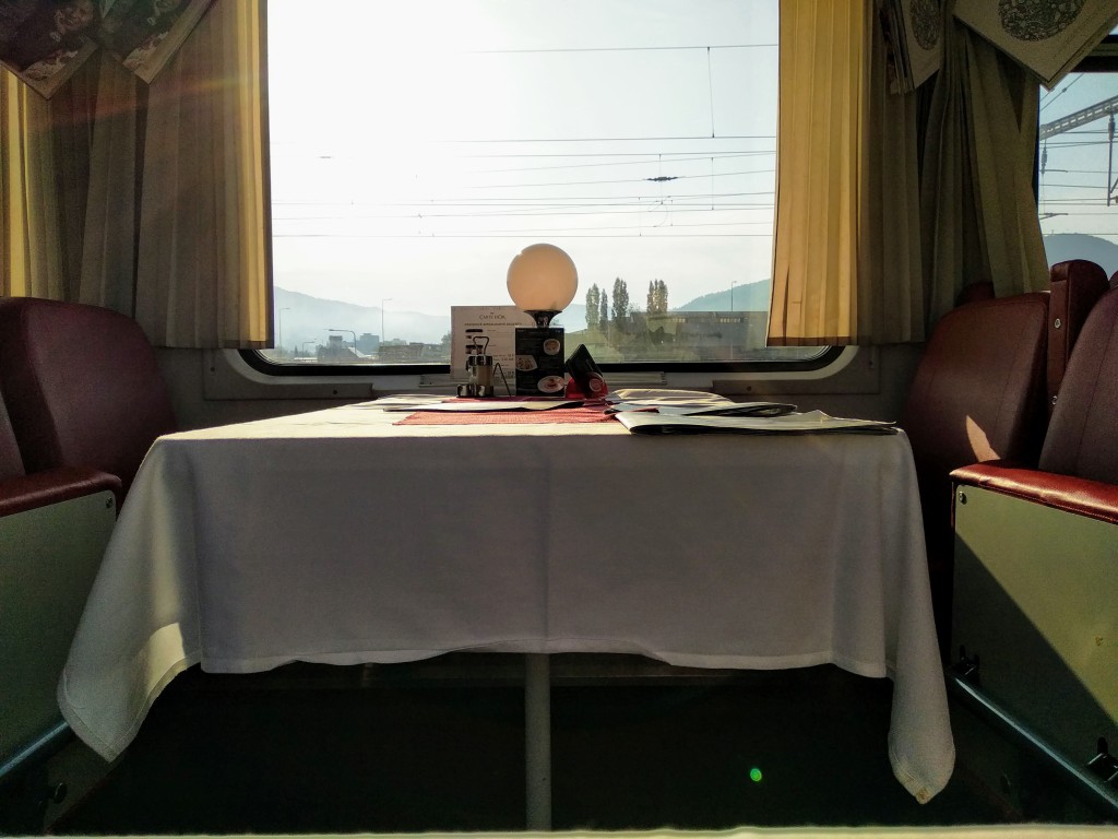 Interior view of a train dining car with a white tablecloth covering a table set with a lamp, menus, and condiments, overlooking a landscape through the train window.