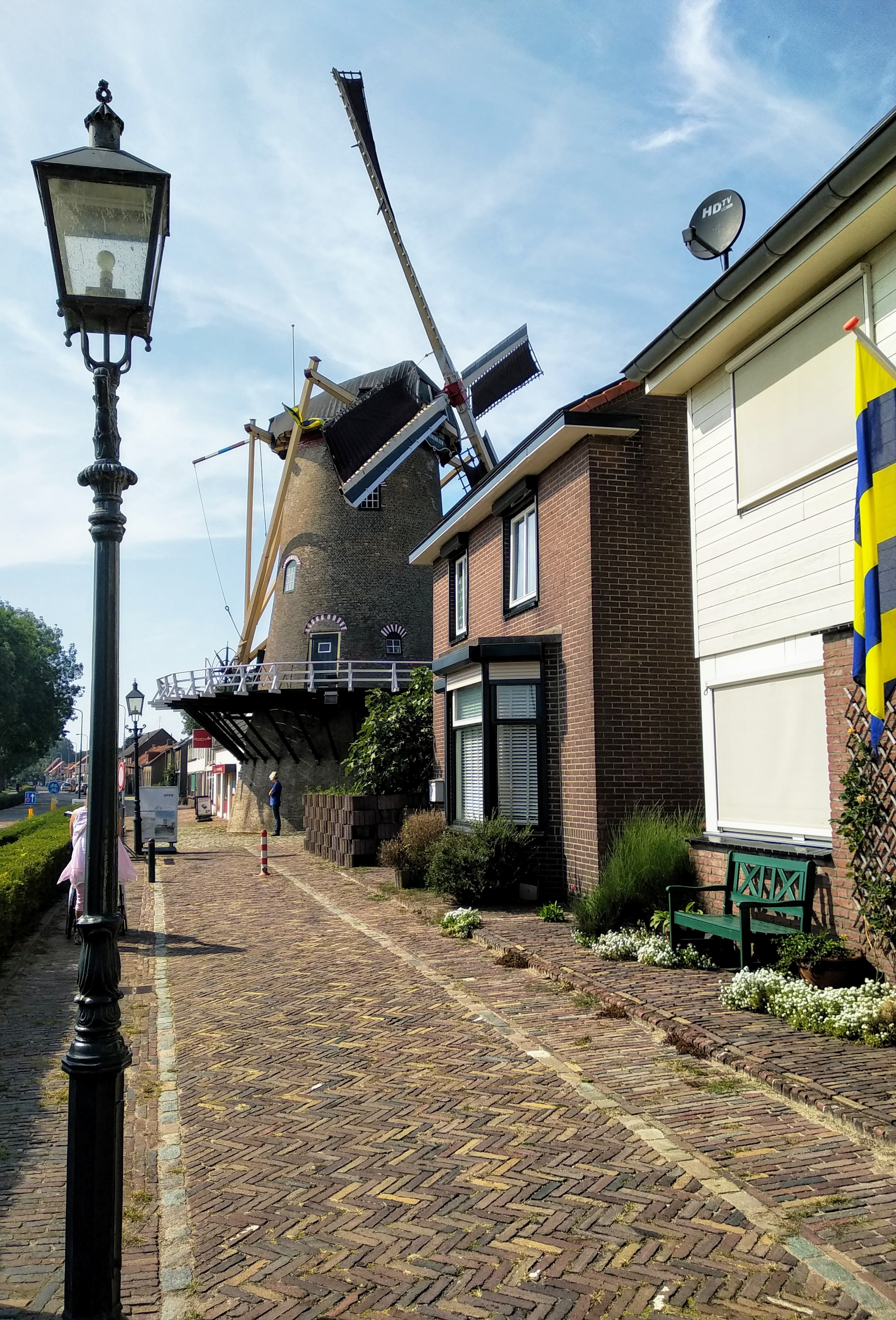 A brick-built Dutch windmill with extended sails alongside a brick building and sidewalk, with a black lamppost in the foreground, and a sunny blue sky background.