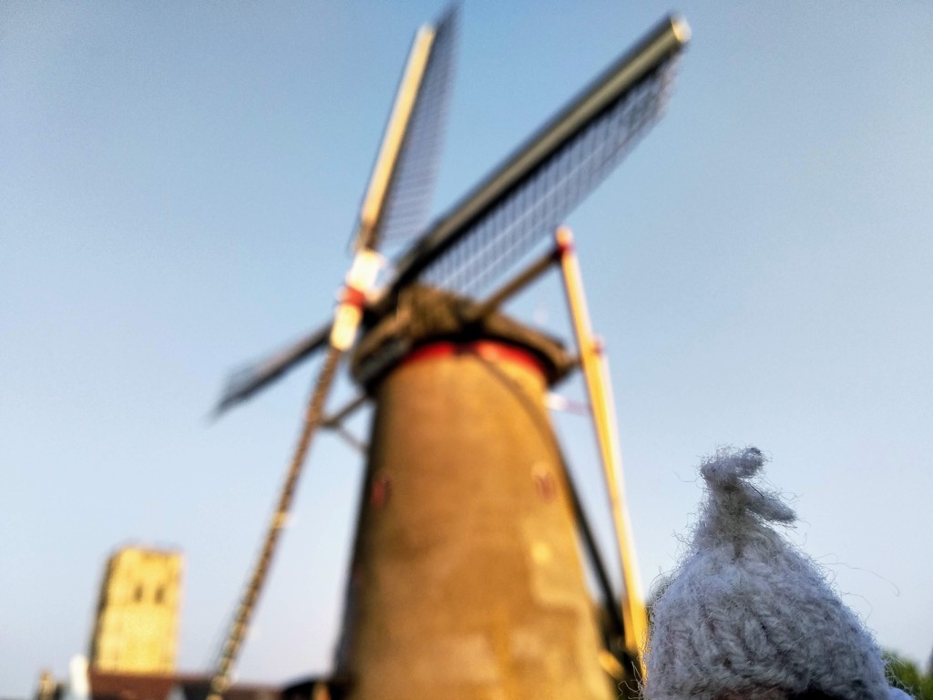 A tan Dutch windmill with black vanes against a light blue sky is out of focus, because there is a finger puppet in the foreground.