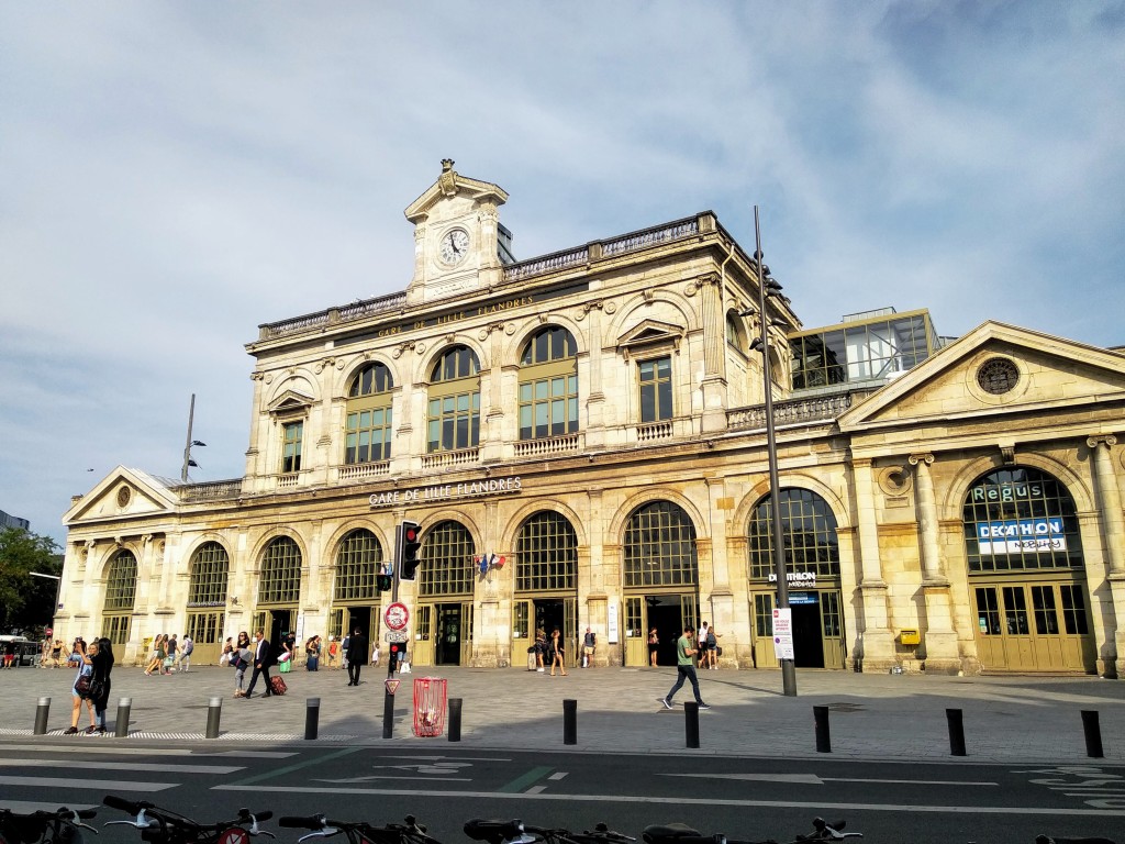 Exterior view of the Gare de Lille-Flandres, a classical building with arched entrances and a clock tower, with pedestrians entering and exiting the station.